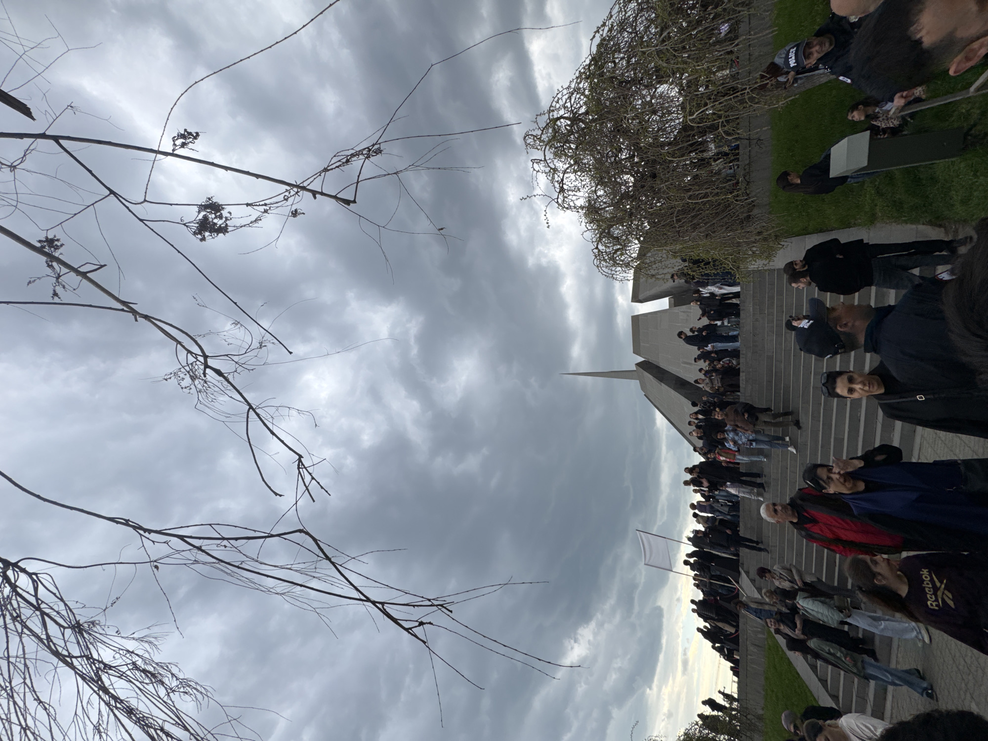 The rotated image captures an outdoor gathering of a large crowd of people, set against an overcast sky. In the immediate foreground, dark, bare tree branches with some dried foliage extend across the frame, partially obscuring the cloudy, grey sky.
The main focus is a diverse crowd of individuals, varying in age and attire, standing on wide concrete steps that ascend towards a distinctive, angular concrete monument. This structure features sharp lines and a prominent, slender spire pointing skyward. The monument's architectural style suggests a modern memorial or public landmark.
To the right of the steps, patches of green grass are visible. The atmosphere appears solemn, with many people looking forward or engaging with the event. One person in the crowd holds a plain white flag on a pole. The weather is characterized by heavy, low-hanging grey clouds, indicating an overcast day.
Notable visible text includes "POLICE" on a dark vest worn by an individual situated on the grassy area, suggesting a security presence. Another person's sweatshirt bears the brand name "Reebok." Given the location context of Yerevan, Armenia, the scene likely depicts a commemorative event or public assembly at a significant national monument.