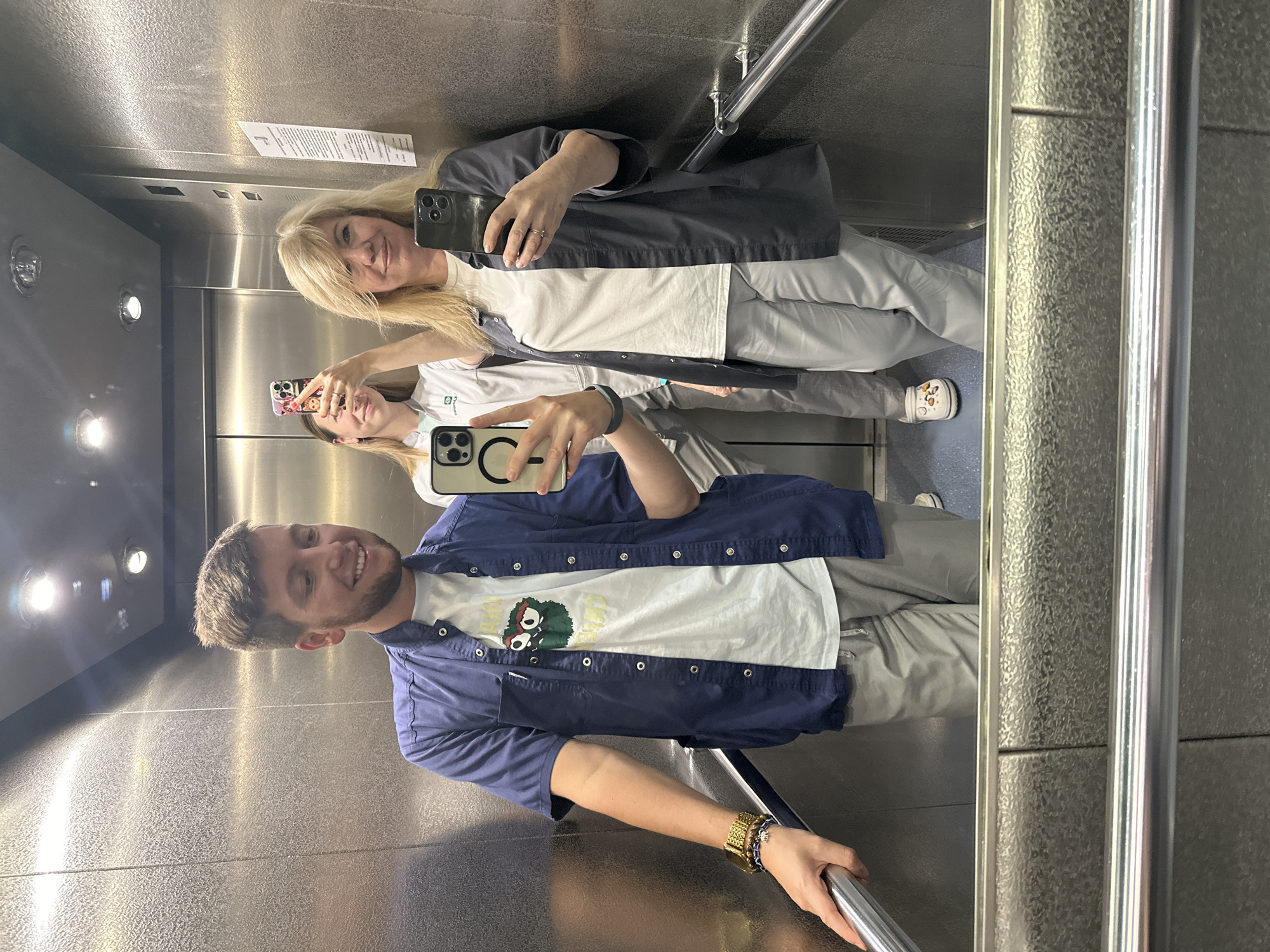 Three individuals are taking a mirror selfie inside an elevator. On the left, a male with short brown hair and a beard smiles and leans on the elevator handrail, wearing a blue short-sleeved button-up shirt over a white t-shirt with a graphic print, and grey pants. In the middle, a female with blonde hair, wearing a white lab coat over light grey pants, holds up a phone to take a selfie. On the right, another female with blonde hair, also wearing a light grey button-up jacket over a white t-shirt and light grey pants, holds up a phone to take a selfie. The elevator interior has a metallic finish and fluorescent lighting. The image is reflective, showing the floor and handrails. There is no indication of the city of Moscow, Russia.