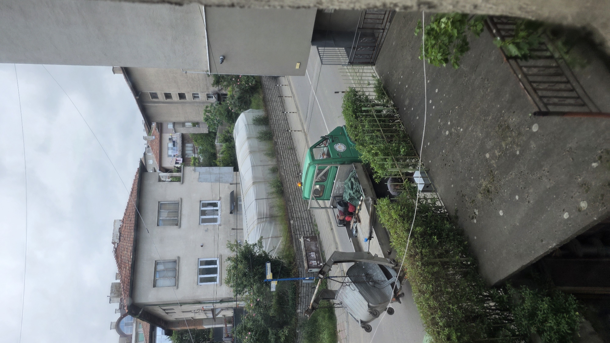 The image shows a view of a street in Blagoevgrad, Bulgaria, taken from an elevated position, likely a balcony or window. The scene depicts a green utility truck lifting a large, gray metal waste container. The truck is parked on the side of the road, near a sidewalk bordered by a hedge. A building with white-framed windows and a tiled roof is visible on the left side of the street. In the background, there's a white greenhouse-like structure and other buildings. The sky is overcast.