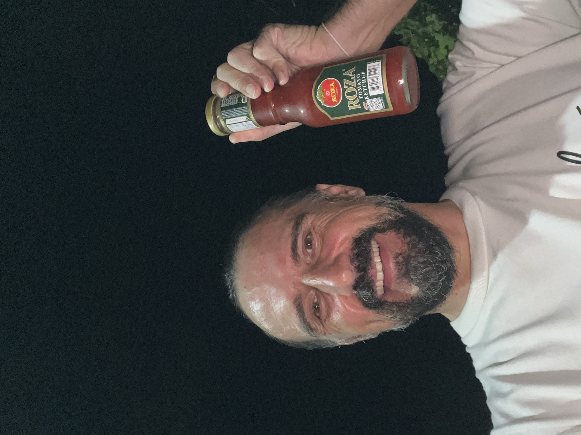 A male subject is depicted in a medium shot, smiling directly at the camera while holding a glass bottle of 'ROZA TOMATO KETCHUP' in his right hand. The subject has a dark beard and mustache, with dark hair pulled back, and is wearing a white t-shirt. The ketchup bottle has a green label with the brand name 'ROZA' and a red tomato graphic, topped with a gold-colored cap. The scene is illuminated by a direct light source against a predominantly dark background, indicating nighttime conditions. Minimal dark green foliage is discernible on the right side of the frame. This image was captured in บ้านบางรักษ์, Thailand.