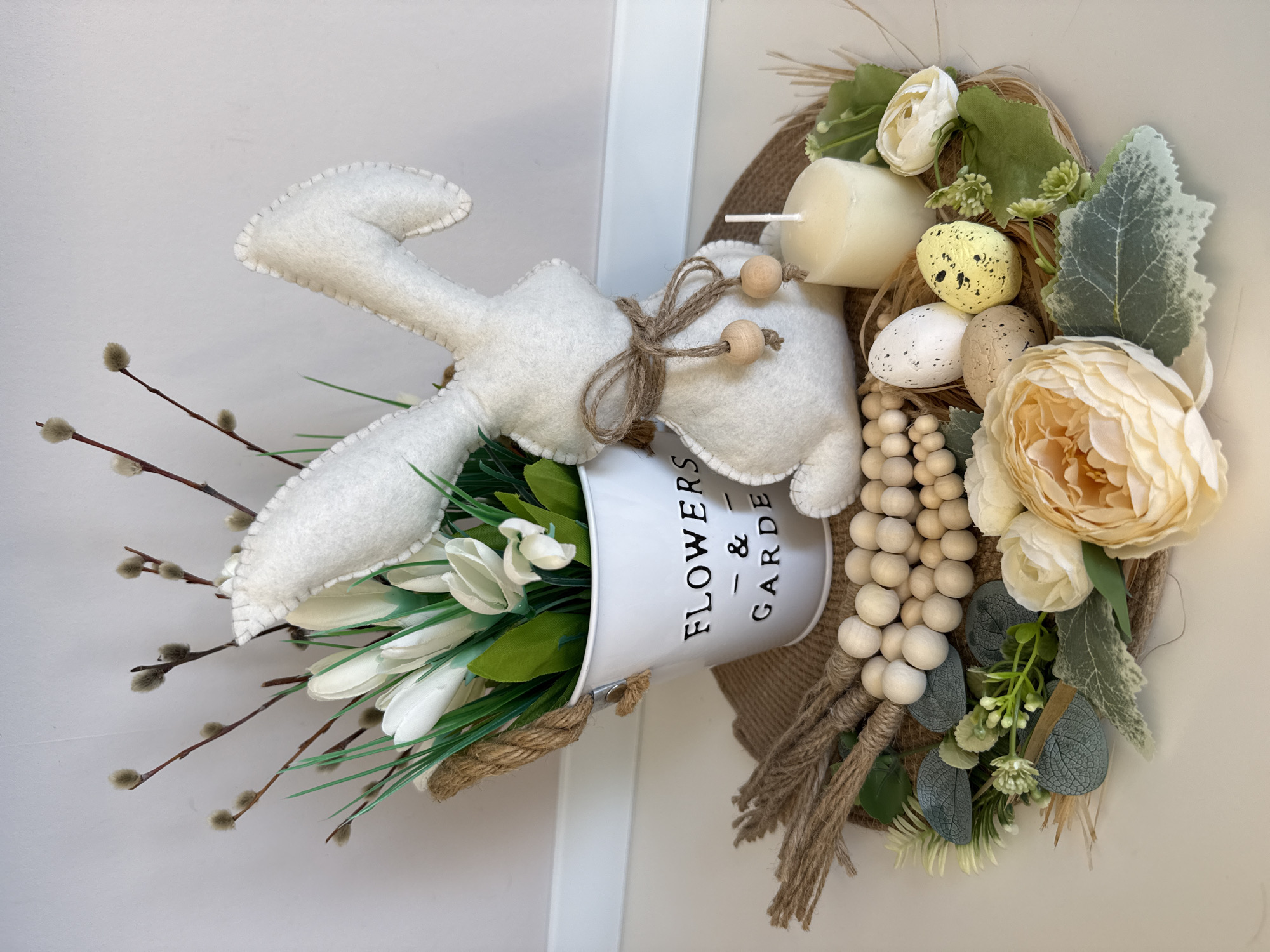 The image shows a decorative Easter wreath hanging on a white surface, likely a door or wall. The wreath is constructed with a burlap base. A white metal bucket labeled "FLOWERS - & - GARDEN" holds faux white tulips and pussy willow branches. A felt bunny with a twine bow is positioned to appear as if it is coming out of the bucket. On the right side of the wreath are faux flowers in peach and white, along with speckled eggs and wooden bead garlands. There is also a short white candle incorporated into the design. The scene depicts a handcrafted, holiday-themed decoration. The image does not show the location in the city of Votkinsk, Russia.