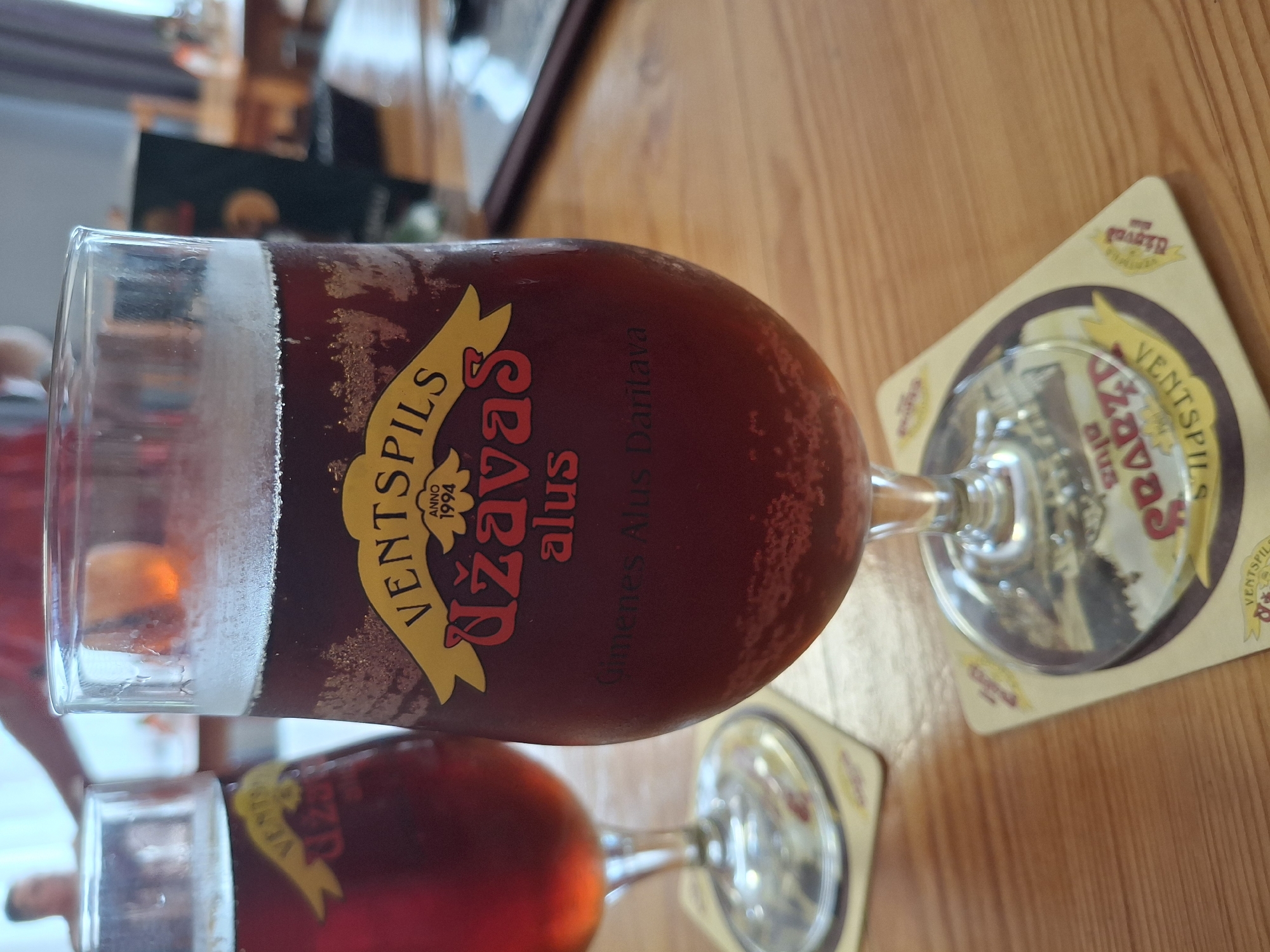 A medium shot captures two glasses of dark beer on a wooden table. The glass in the foreground, which is larger and in focus, displays a yellow ribbon-like label with "VENTSPILS" written in black at the top, followed by "ANNO 1994" and "UŽAVAS" in red script. Below this, "alus" is written in smaller red script, and then "Ģimenes Alus Darītava" in grey. The beer has a frothy head and condensation on the glass. The second glass, out of focus to the left, shows a similar label partially obscured. Coasters with similar branding are beneath each glass. The background is blurred, suggesting an indoor setting with people and furniture, possibly a pub or restaurant in Lēdmane, Latvia, given the branding on the glasses.