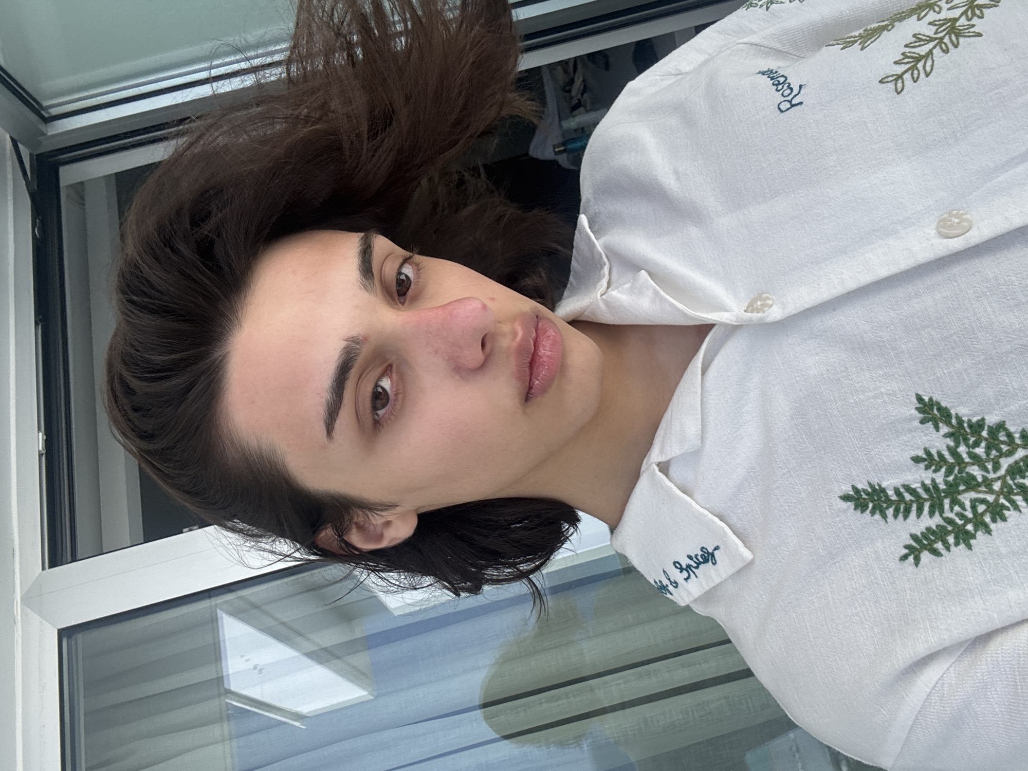 A close-up portrait, rotated 90 degrees clockwise, features a young person with dark brown hair and fair skin looking directly into the camera. Their head is positioned on the left side of the frame. They have dark eyes, well-defined eyebrows, and full lips with a subtle sheen. A slight pinkish flush is visible on their nose and cheeks.
The person is wearing a white button-up shirt made of a linen-like fabric. The shirt features green embroidered patterns resembling leafy sprigs. Visible text embroidered on the shirt includes "Rosemary" near one of the leafy designs and "Salt & Spice" on a smaller part of the shirt, possibly a cuff or collar. Several white buttons are also visible on the front of the shirt.
The background consists primarily of a large window or glass door with white framing, through which bright daylight is visible. Reflections on the glass suggest interior elements like blinds or distant structures. The setting appears to be indoors, likely in a well-lit space adjacent to the outdoors, consistent with a sunny climate such as Ayia Napa, Cyprus. The person maintains a neutral and calm expression.
