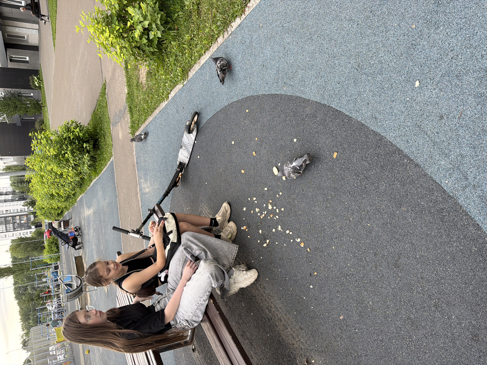 Two young females are seated on a wooden bench in a modern urban playground. The female on the left is older, wearing a black t-shirt and light blue jeans, with long brown hair and glasses. The female on the right is younger, wearing a black sleeveless top and a dark skirt, with her hair tied up. They are looking in different directions and appear relaxed. In front of them, on the ground, are two kick scooters and scattered pieces of food, likely bread crumbs. Two pigeons are visible on the ground, pecking at the food. The playground surface is a mix of dark grey and light blue rubberized material, typical of modern urban play areas. In the background, there is a children's play structure, lush green bushes, and a multi-story building, suggesting an urban residential area. The presence of a stroller in the background further indicates a family-friendly environment. The overall scene is consistent with a public park or a courtyard in a residential complex in Kazan, Russia, during daytime, with overcast skies.