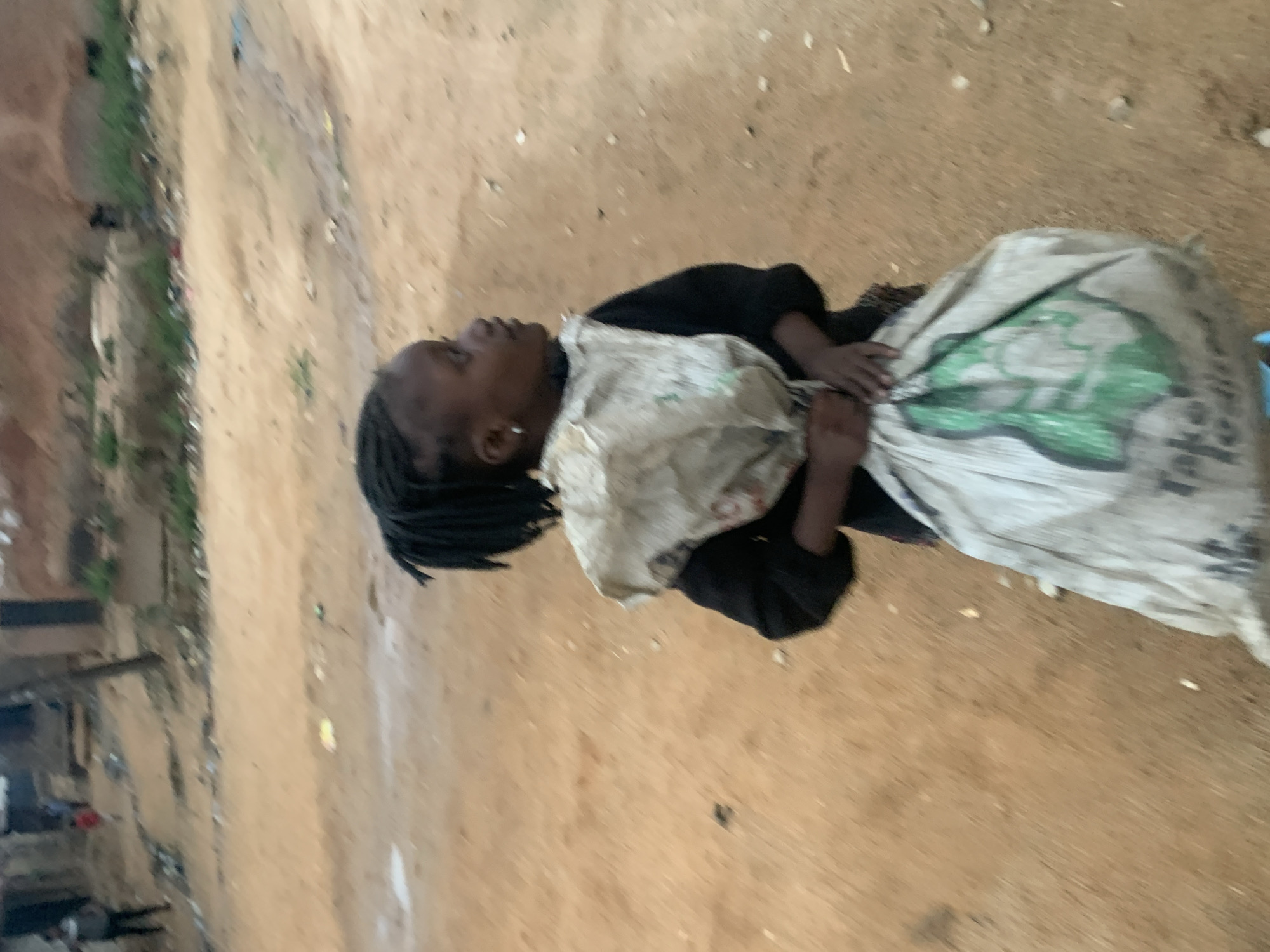 A child lies on a dirt road, clutching a large burlap sack. The child wears a dark top and appears to have braided hair. Debris is visible along the roadside. The location is Jos, Nigeria.