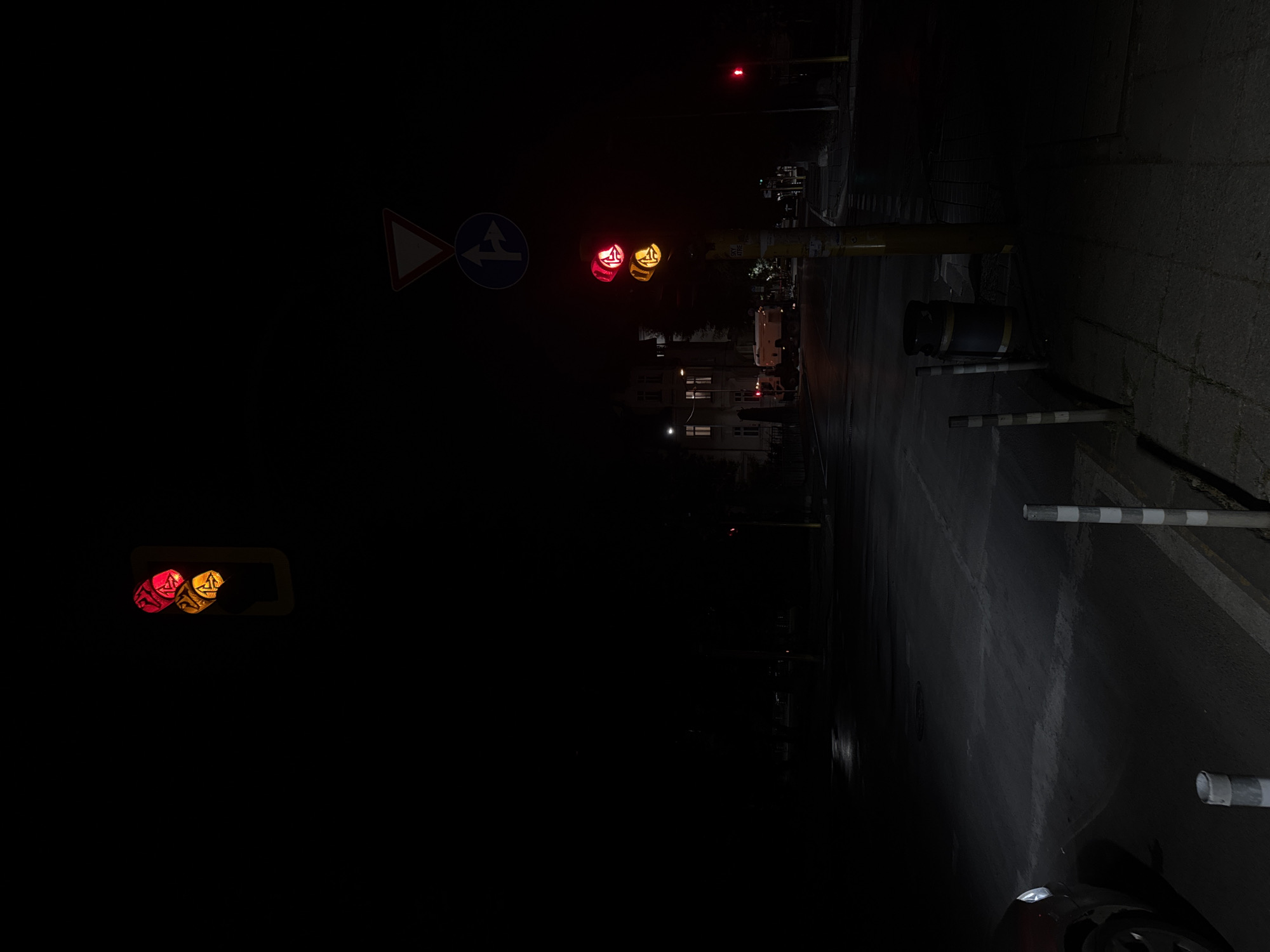 The image captures a nighttime scene at an intersection in Sofia, Bulgaria. A traffic light in the foreground shows a red light at the top and a yellow light below, indicating a warning or an approaching change in signal. Above this traffic light, a triangular yield sign is visible, followed by a blue circular sign with a white arrow pointing left and another arrow pointing right, indicating allowed turns. To the right of the main traffic light, another pole with a single red traffic light is visible in the distance, suggesting traffic flow in another direction.
The intersection appears wet, possibly from recent rain, reflecting the ambient light. Streetlights and lights from buildings in the background illuminate the scene, casting long shadows. A red vehicle with its headlights on is partially visible on the left side of the image, suggesting it is stopped at or approaching the intersection. In the mid-ground, a large building with illuminated windows is present, and a white truck is parked near it.
On the sidewalk in the foreground, a trash bin is situated next to the traffic light pole. Several striped bollards are placed along the edge of the sidewalk, separating it from the road. The overall impression is one of a quiet, dimly lit urban intersection at night, with functioning traffic signals and minimal activity.