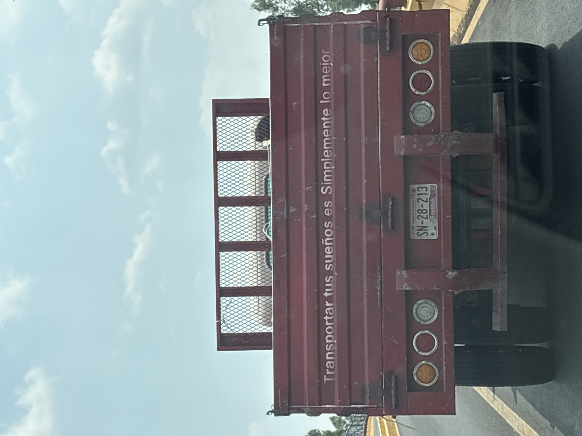 The image shows the back of a red flatbed truck with the text "Transportar tus sueños es Simplemente lo mejor" painted on its side. The truck has a mesh cage behind the cab and standard taillights. The license plate, partially visible, reads "SN-28-213" and identifies the vehicle as being from Puebla, Mexico. The image appears to have been taken from another vehicle while in transit. The sky is visible in the background, suggesting it is daytime.