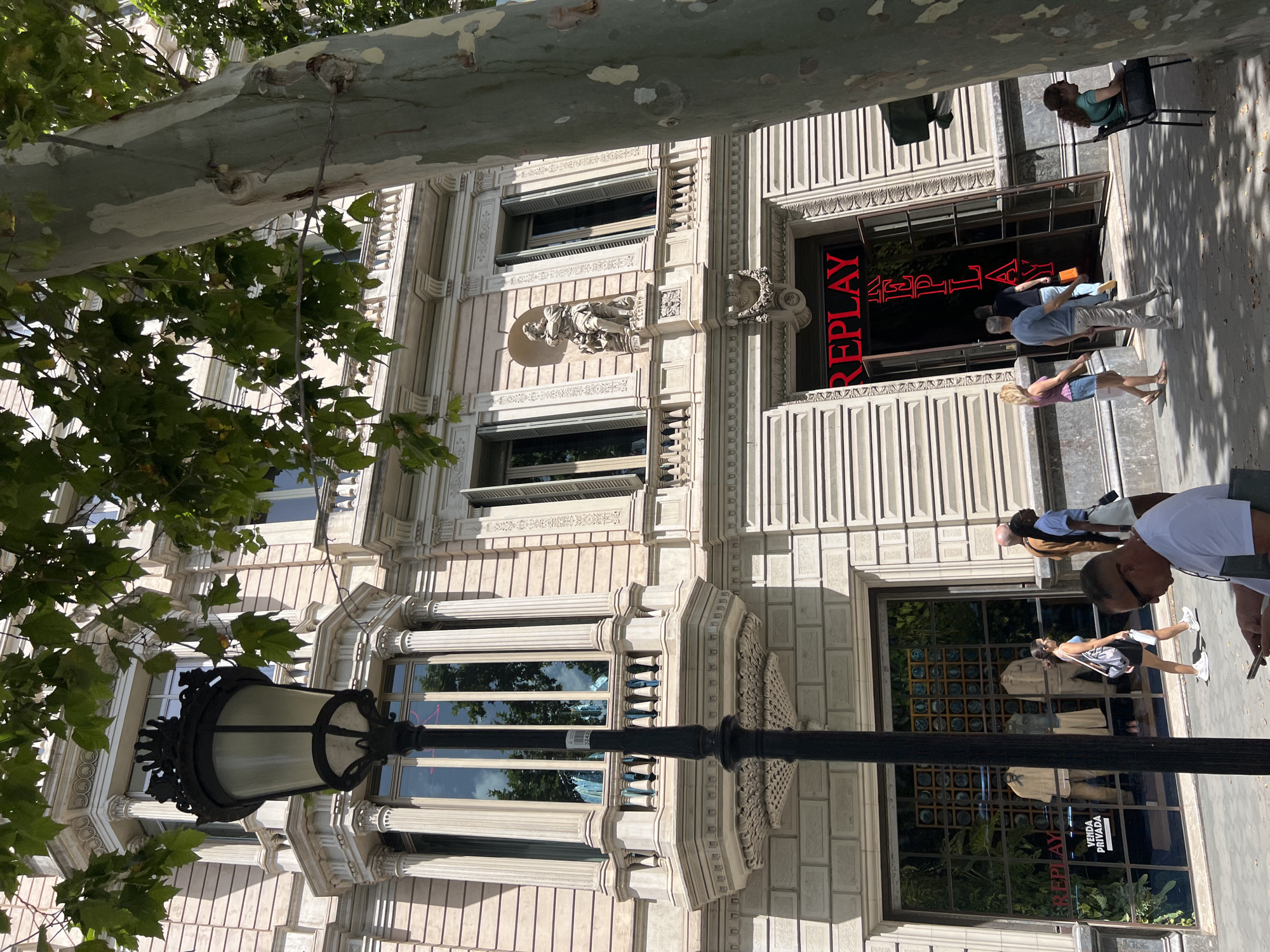 The image captures a street scene in Barcelona, Spain, featuring the facade of a building with architectural details and a Replay store. In the foreground, a black lamppost with intricate scrollwork stands prominently. Partially obscuring the upper portion of the building and lamppost is the trunk and foliage of a large tree.
The building itself is light-colored, with a classical architectural style. It has multiple stories with arched windows, balconies, and decorative moldings. One prominent bay window features tall columns and a detailed balcony. A sculpture of a person is visible on the second story, above an open doorway.
The ground floor houses a Replay store, indicated by large red neon lettering above the entrance. Two glass doors provide access to the store. To the left of the Replay entrance, another large storefront displays clothing items and greenery.
Several people are present on the sidewalk. A woman in a blue top and white shorts walks past the clothing store. Further down, a man in a light blue shirt and beige pants walks towards the Replay store, followed by a person in black. To the far right, a woman with long brown hair sits in a black chair, facing away from the camera. Another person in a white t-shirt and sunglasses is seated in the foreground, looking down at a phone.
The street appears to be paved, and shadows from the tree and building suggest it is a sunny day. The overall scene depicts a typical urban environment with commercial activity and pedestrian presence.