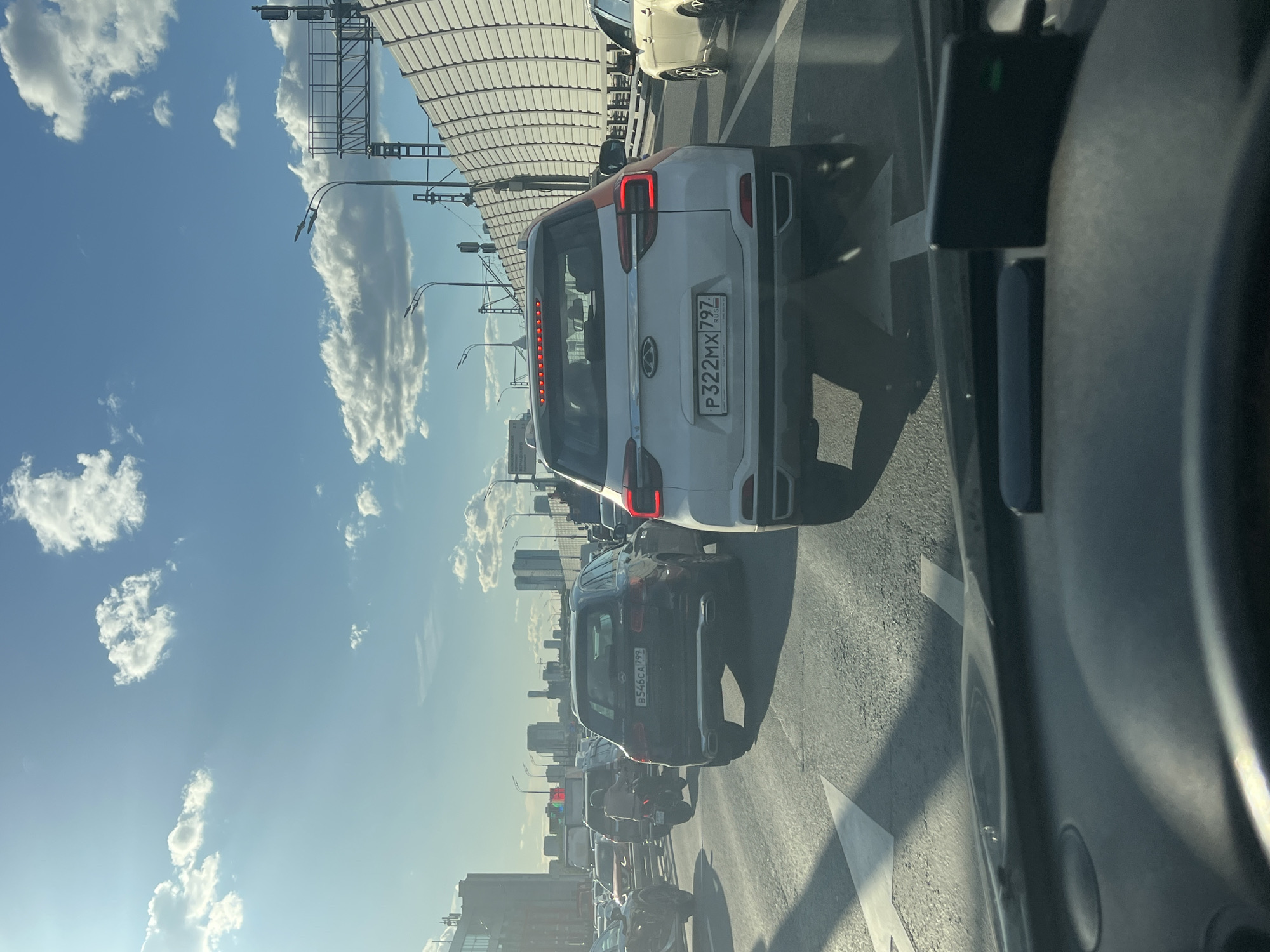 The image depicts a traffic jam on a sunny day in Moscow, Russia, likely during daylight hours. The perspective is from inside a vehicle, looking forward. A white car with an orange stripe on its rear and a license plate reading "P322MX 797" is prominently visible in the foreground. Behind it, a dark SUV with a license plate "B546CA 799" and further vehicles are seen, all stationary in traffic. The background shows a modern cityscape with tall buildings and a distinctive overpass structure with a curved, ribbed canopy. The sky is blue with scattered white clouds. The overall impression is one of congestion in an urban environment under clear skies. There is no indication of specific emotions or sensitive content.