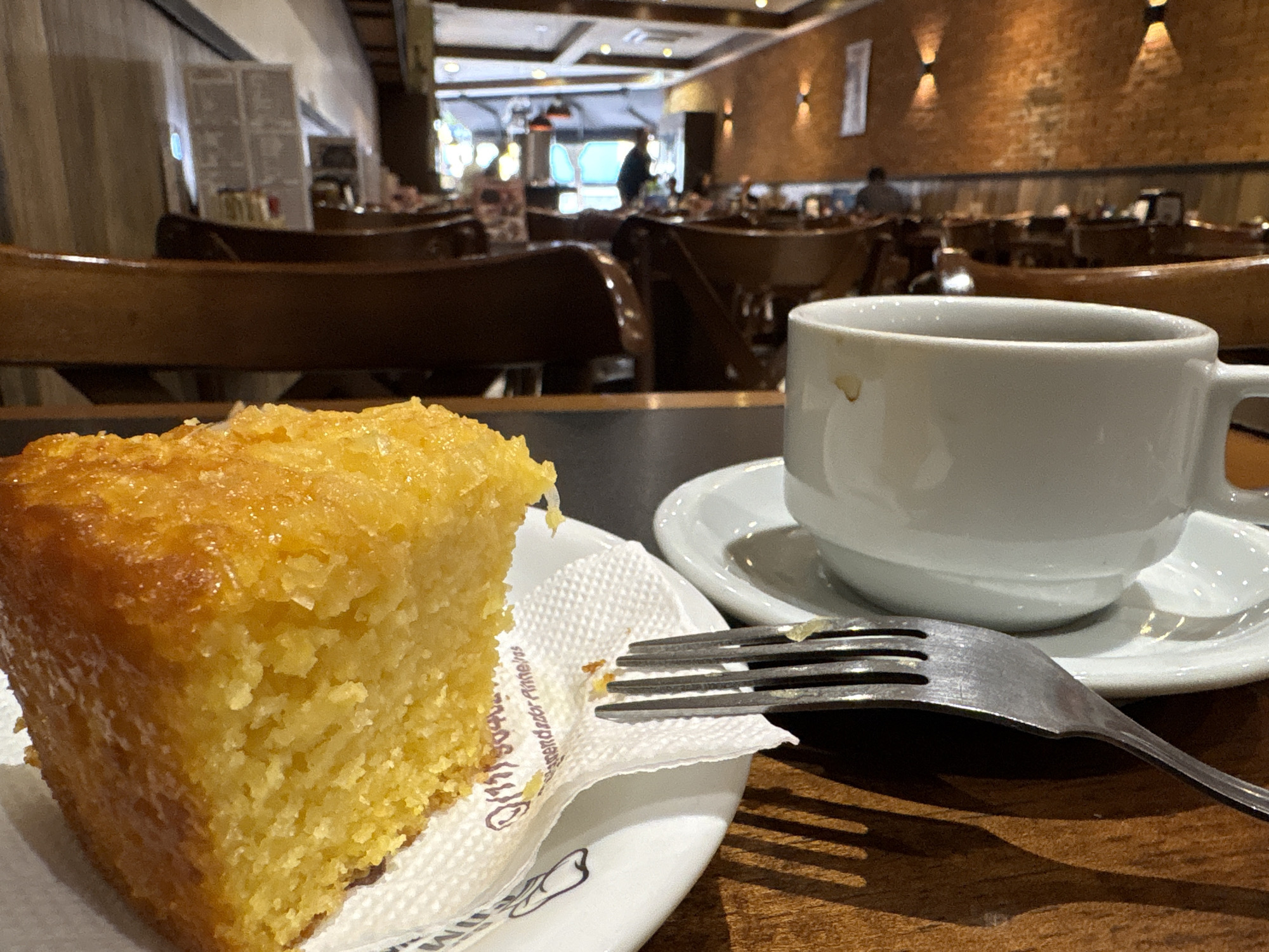 A close-up shot shows a slice of corn cake and a white coffee cup on a dark wooden table in a cafe. The cake is on a white plate with a napkin, and a fork is resting on the napkin. The background is blurred, showing tables and chairs in what appears to be a cafe or restaurant setting. The cafe's interior is decorated with exposed brick walls and wooden furniture. The lighting suggests it is daytime or early evening. The image is focused on the food and drink, with the background providing context for the location.