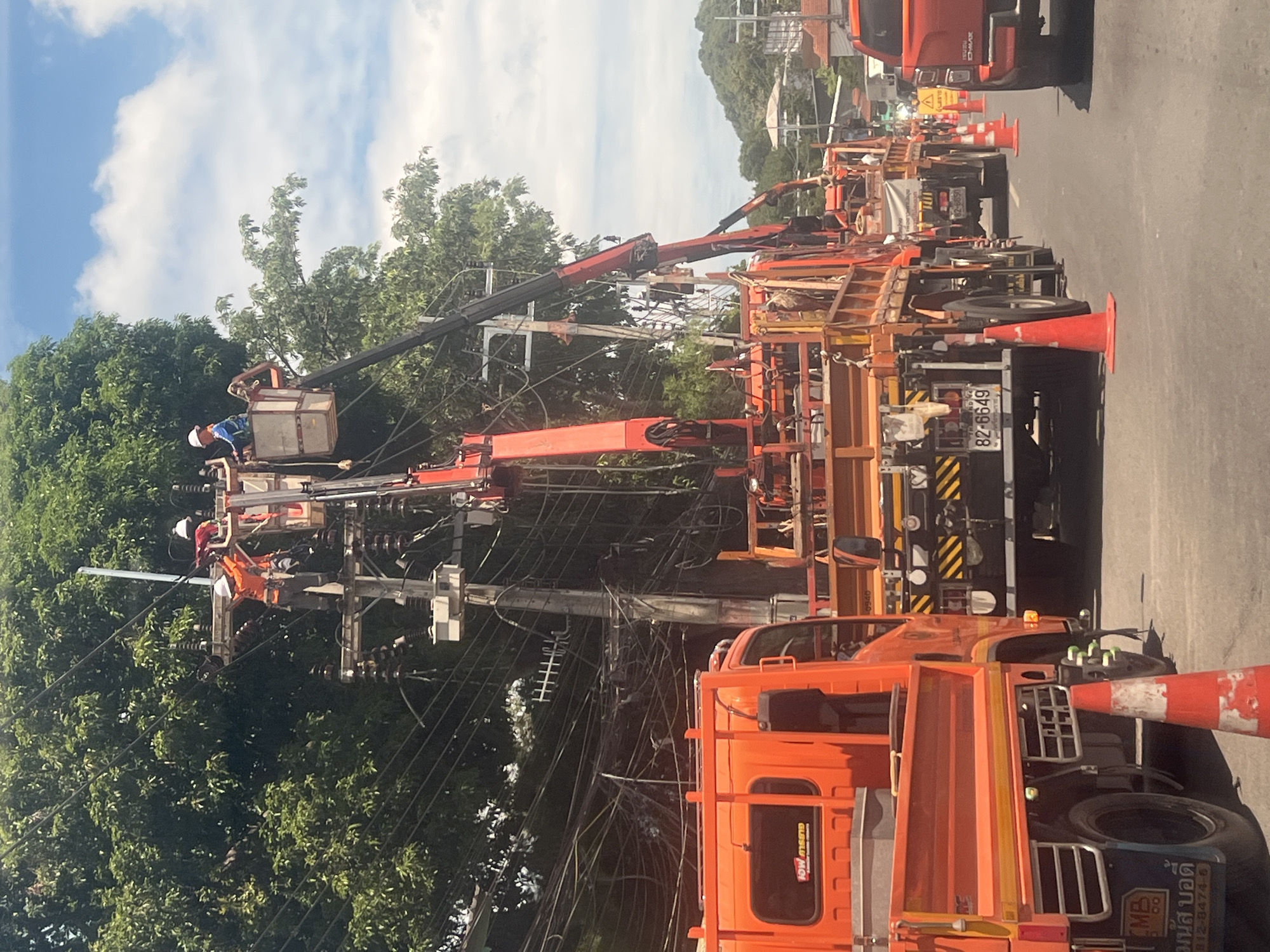 In the city of Ko Samui District, Thailand, two utility workers are performing maintenance on overhead electrical lines from elevated work platforms. Both workers are wearing hard hats and safety vests. The first worker, in an orange vest, is positioned higher on a utility pole, appearing to adjust equipment. The second worker, in a blue and white vest, is in a basket lift attached to a crane, also working on the lines. Several large orange utility trucks are parked on the road, with their cranes extended to support the workers. The area is characterized by dense foliage from large trees lining the street, and a clear sky with scattered clouds. The ground is asphalt, and orange traffic cones are placed along the road. The scene suggests ongoing electrical infrastructure work.