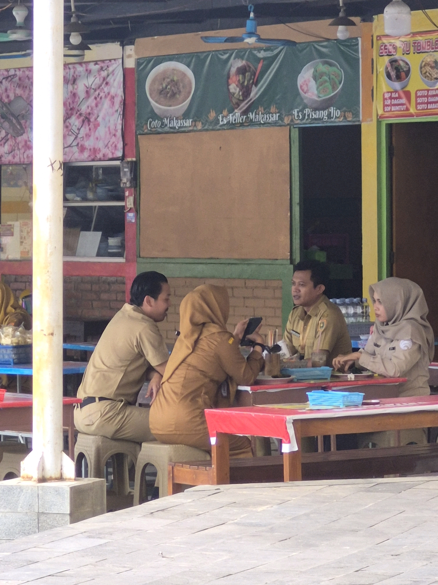 This image captures an outdoor dining scene at what appears to be a food stall or casual restaurant, possibly located in the city of Purworejo, Indonesia. Four individuals, all dressed in tan or khaki uniforms which suggest they might be government employees or civil servants, are seated around a red-covered table. A man in the center is engaged in conversation, gesturing with his right hand. To his right, a woman in a beige hijab is looking down at the table, while a woman in a similar hijab to his left is looking at her phone. Another man, on the far left of the table, is also seated and looking away from the camera. The table is laden with several items, including blue plastic containers, glasses, and possibly food or drinks. In the background, banners displaying food items like "Coto Makassar," "Es Teler Makassar," and "Es Pisang Ijo" are visible, along with another banner with text in Indonesian. The environment suggests a warm, likely tropical, climate with an open-air structure providing shade. The overall impression is a casual break or mealtime for uniformed individuals.