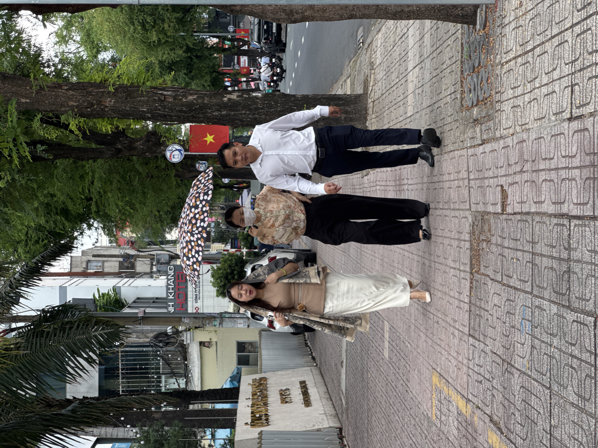Three people walk on a sidewalk in front of a street. The person on the left, a woman, wears a light brown shirt, a white skirt, and a patterned scarf around her shoulders. She is holding a phone in her right hand. The person in the middle, a woman, wears a patterned shirt, black pants, and a face mask. She is holding a closed umbrella over her head. The person on the right, a man, wears a white button-down shirt and dark pants. He is walking. The sidewalk is paved with textured concrete tiles. In the background, there are buildings, trees, and a street with traffic. A Vietnamese flag with a yellow star is visible on a pole behind the middle person. The scene appears to be in an urban environment.