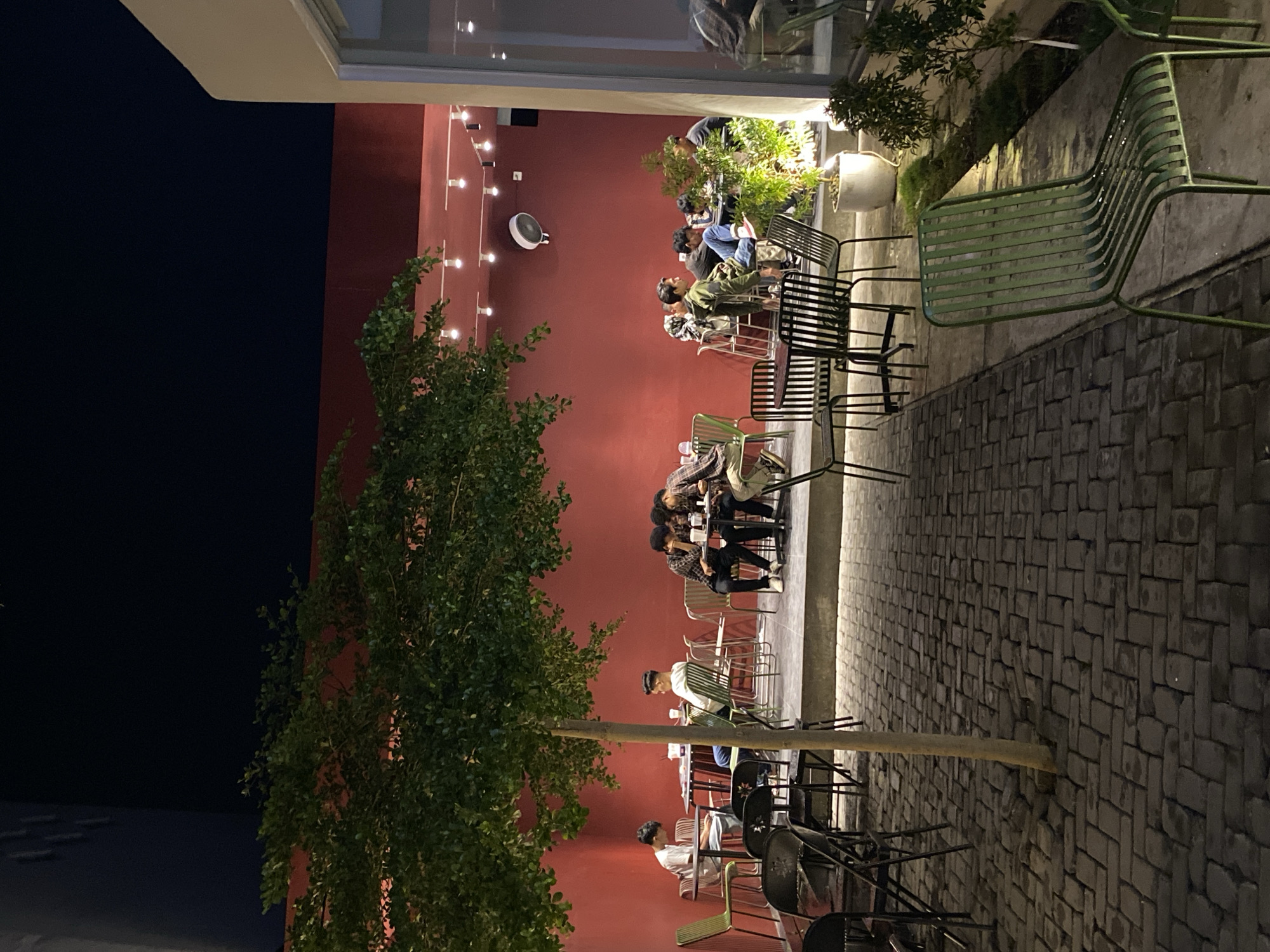 This image depicts an outdoor evening scene at a cafe or restaurant patio, likely in Lhokseumawe, Indonesia. The setting features a prominent, vibrant red wall that stretches across the background, illuminated by a series of string lights that create a warm glow. A white circular fan is mounted on the red wall. The ground is paved with dark, rectangular blocks arranged in a pattern, creating a textured surface for the seating area.
Numerous metal chairs and tables are arranged across the patio. Some chairs are a distinct light green, while others are black. Several groups of people, mostly young men, are seated at these tables. They appear to be engaged in social interaction, relaxing, or using their mobile phones, contributing to a casual and communal atmosphere.
Lush green foliage is a significant element of the scene, with a large, leafy tree positioned on the left side, and smaller potted plants or shrubbery situated closer to the red wall, adding natural elements to the urban setting. The dark sky overhead confirms it is nighttime, with all illumination coming from artificial light sources. No visible text is discernible within the frame. The overall impression is one of a lively yet relaxed outdoor social gathering spot after dark.