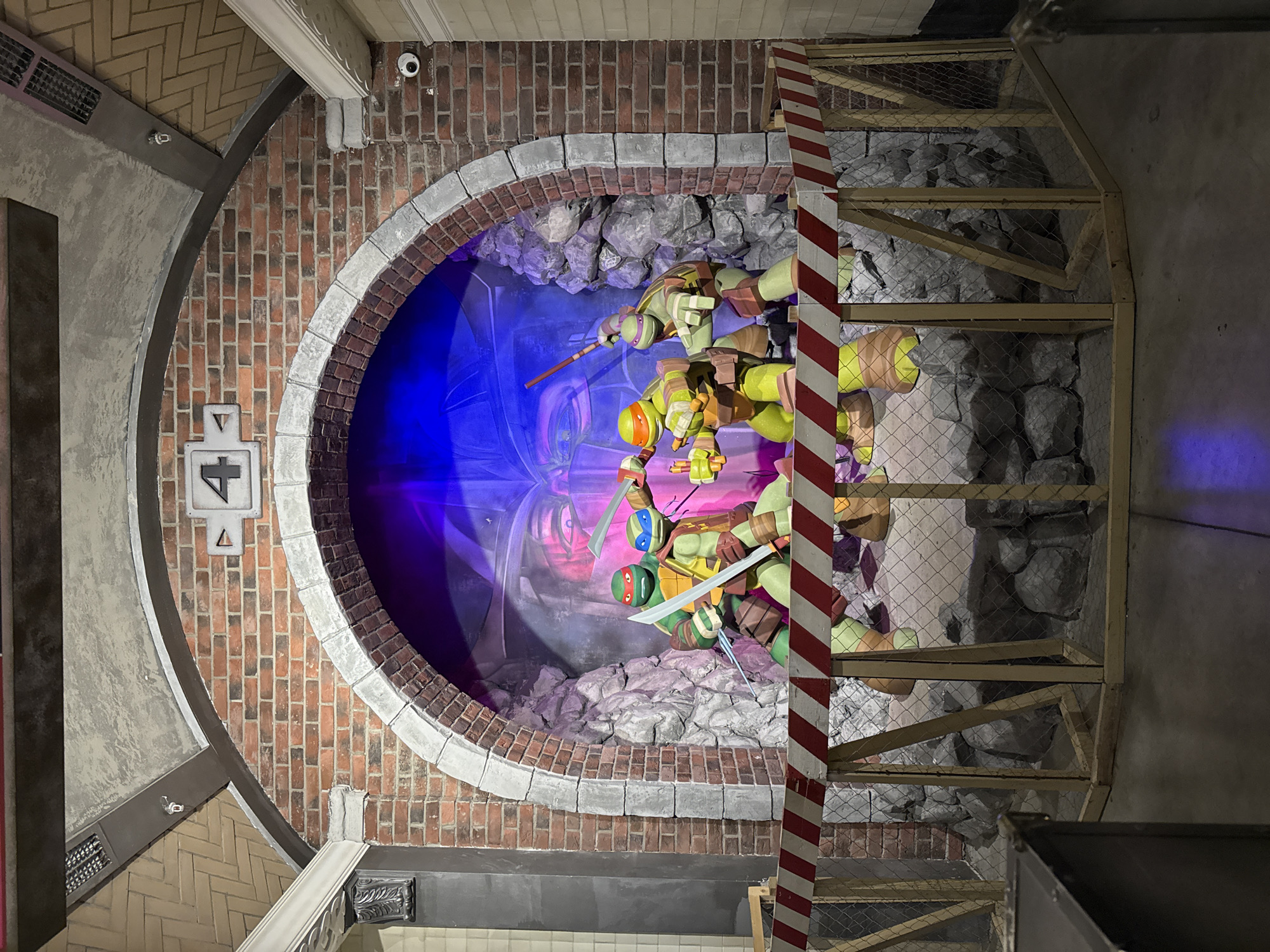 The image shows a Teenage Mutant Ninja Turtles-themed display within an enclosed, constructed environment. The display features representations of the four turtles, along with a backdrop featuring their antagonist, Shredder. The construction of the display incorporates brick and stone-like elements, suggestive of a subterranean or sewer setting. The Turtles appear to be situated on an upper platform, framed by wooden scaffolding and wire mesh.
A sign labeled "4" is visible. Brickwork forms an arch around the display. The display is located in Moscow, Russia.