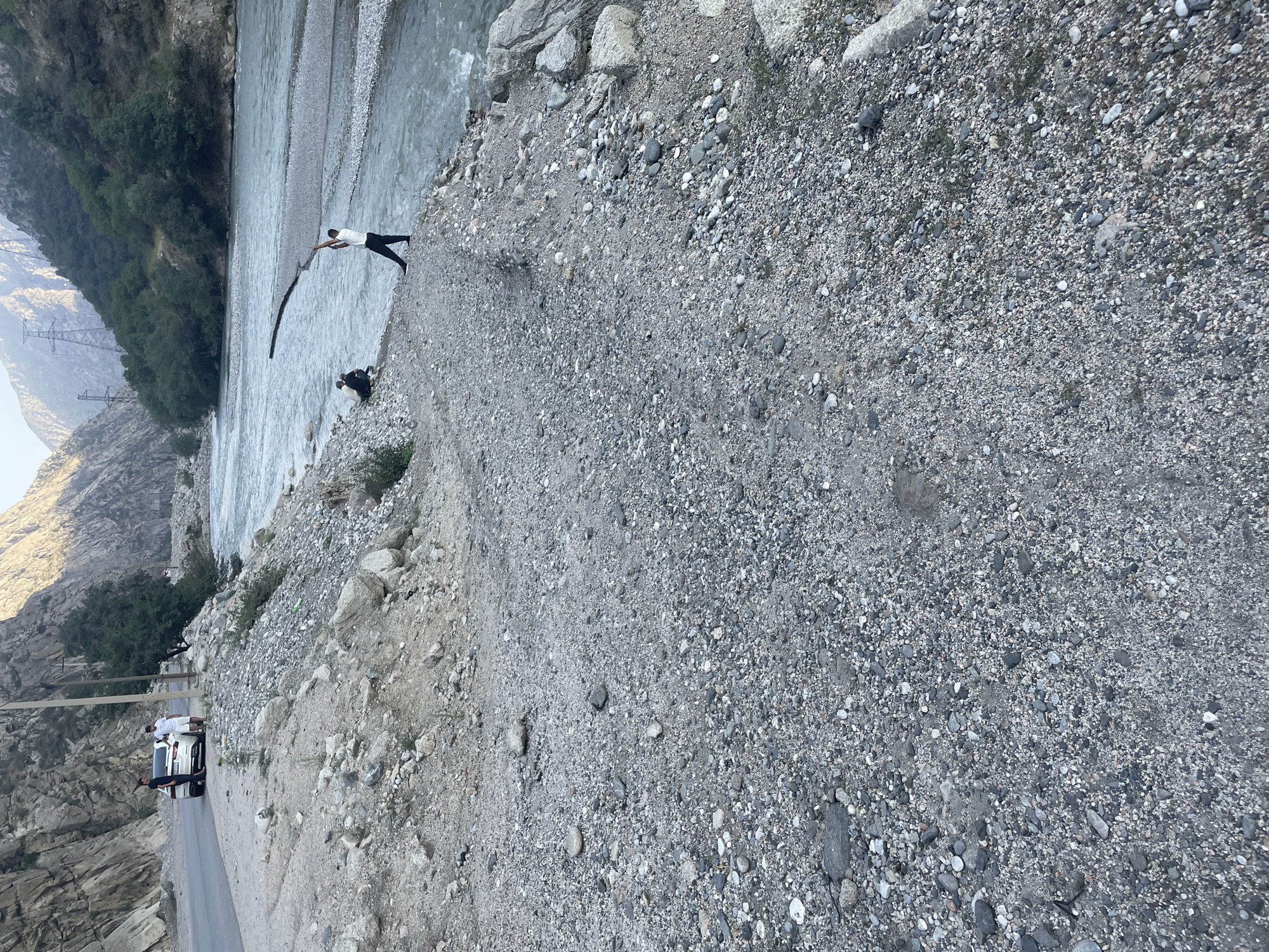 The image captures a view of the river flowing through the city of Khodzha-Obigarm, Tajikistan. In the foreground, a rough, rocky, and gravelly terrain is visible, suggesting a natural, undeveloped area. A paved road runs along the left side of the image, bordered by a rocky embankment. A white car is parked on the side of the road, with two individuals standing near it. Further into the scene, a wide, clear river flows, with a person standing near the riverbank, holding a long object, possibly a stick or pole, and gesturing upwards. Another person is seated on the riverbank, to the left of the standing individual. The background features steep, rocky mountains and lush green trees, with power lines visible on the mountainside, indicating the presence of infrastructure within the natural landscape. The overall scene depicts a semi-urban or peri-urban environment where natural geological features and the river are prominent.