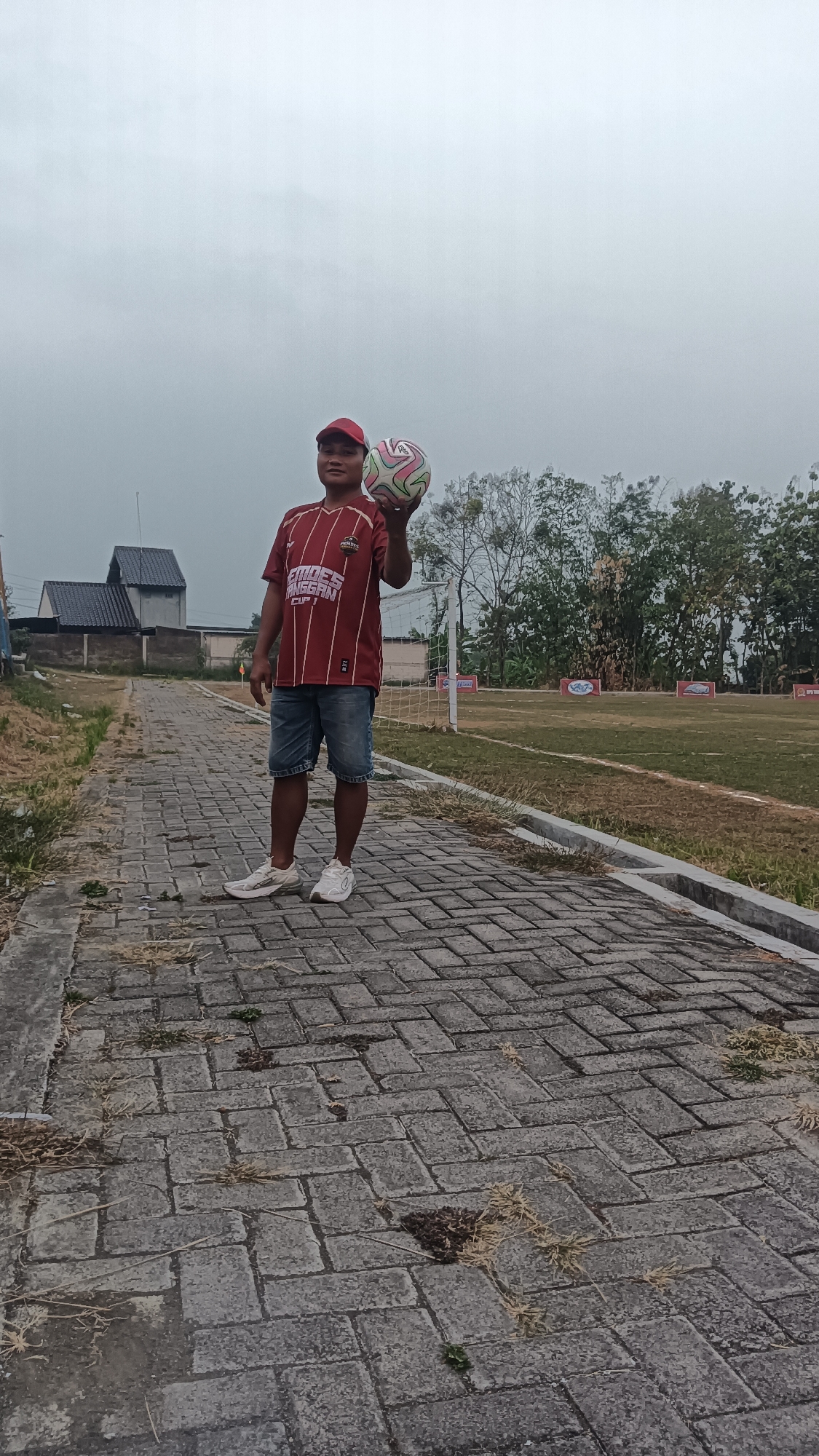 A man stands on a paved walkway in front of a soccer field in Tanggan, Indonesia. The man is wearing a red and white striped t-shirt, blue jean shorts, a red baseball cap, and white sneakers. He is holding a soccer ball in his right hand, which is raised above his head. The soccer field is green with white markings, and there is a soccer goal in the background. The sky is overcast. To the left of the walkway, there are some buildings and trees. The walkway is made of gray pavers and has some dry grass growing between the cracks.