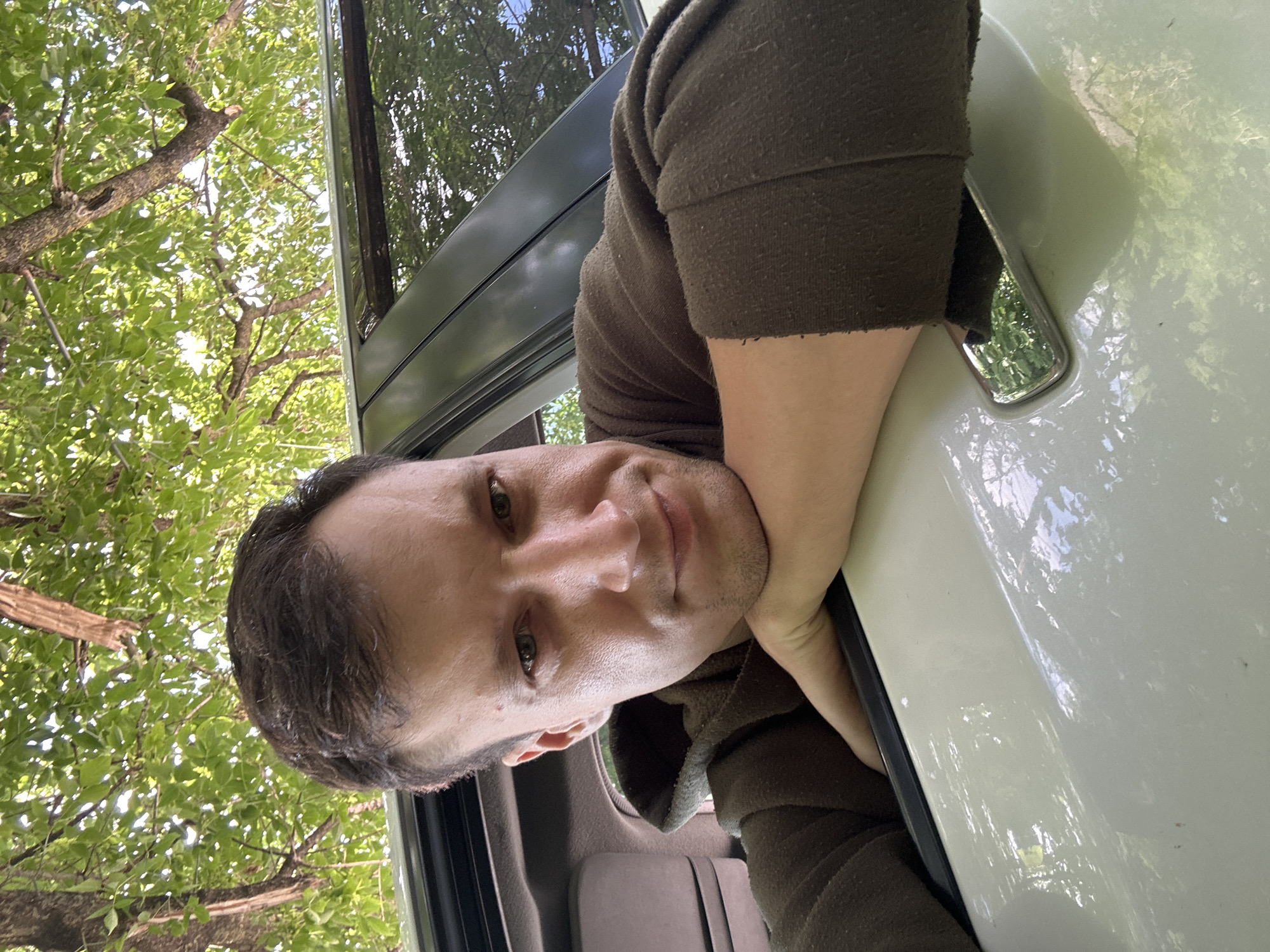 A male is leaning on the door of a white vehicle. He is wearing a brown hoodie. The car is parked outdoors, with trees and foliage visible in the background. The man is looking towards the camera with a slight smile. The image appears to be a selfie, taken from a low angle. The location is Makiv, Ukraine.