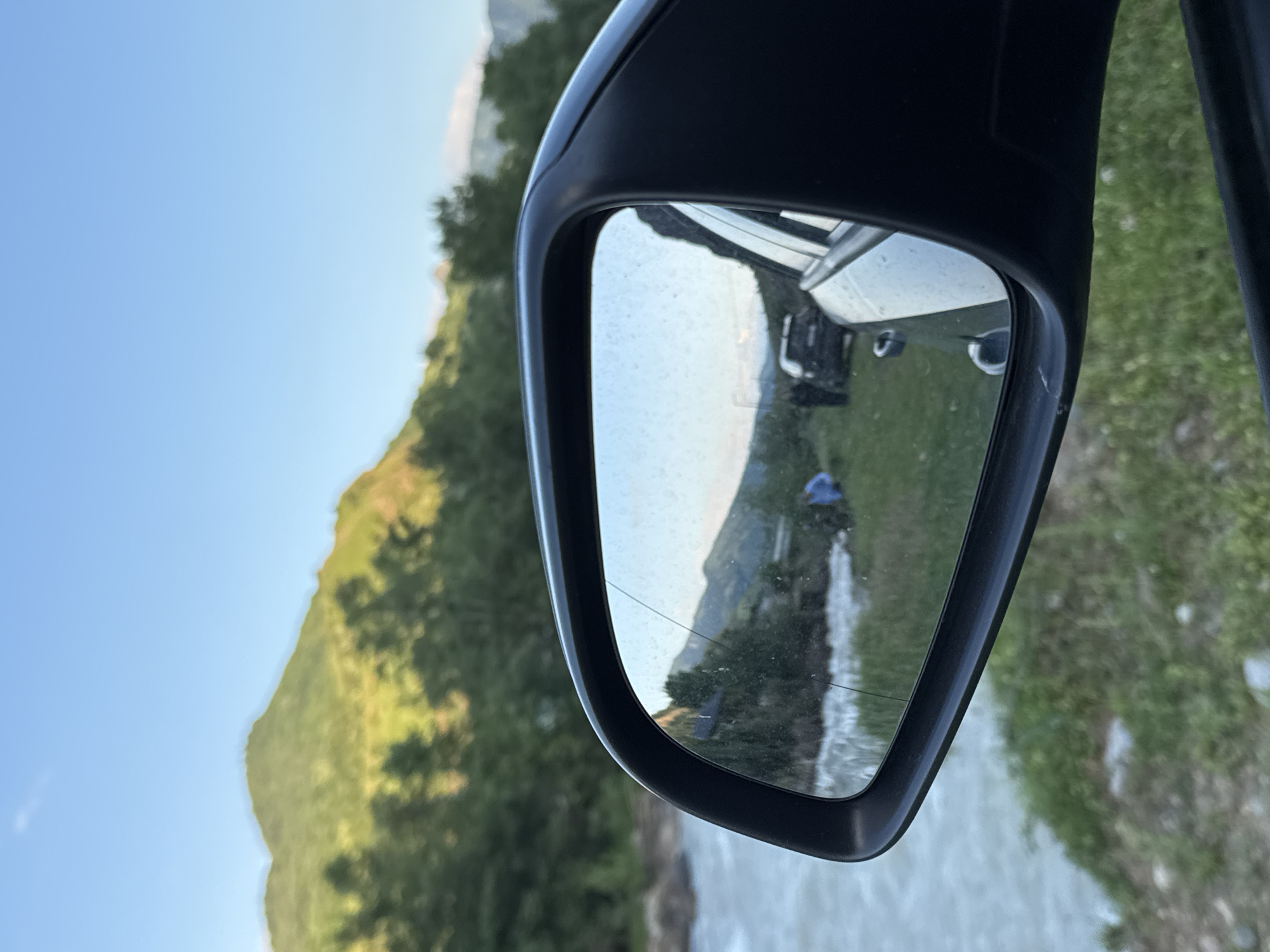 The image shows a view from inside a car, looking at the side mirror, which reflects a scenic landscape. The primary focus is the reflection in the side mirror, which reveals a body of water, possibly a river, flowing to the left of the frame. Beyond the water, there is a grassy embankment with some trees. In the reflection, two figures are visible on the grassy area. One figure, wearing a blue top, is standing near the water's edge. Further back, two dark-colored vehicles are parked. The background of the reflection shows a lush green mountainside under a clear blue sky, with some trees in the middle ground. Outside the mirror, the real-world view shows the side of the car, the grassy ground, and a glimpse of the river to the left. The overall scene suggests an outdoor setting in a rural or natural environment.