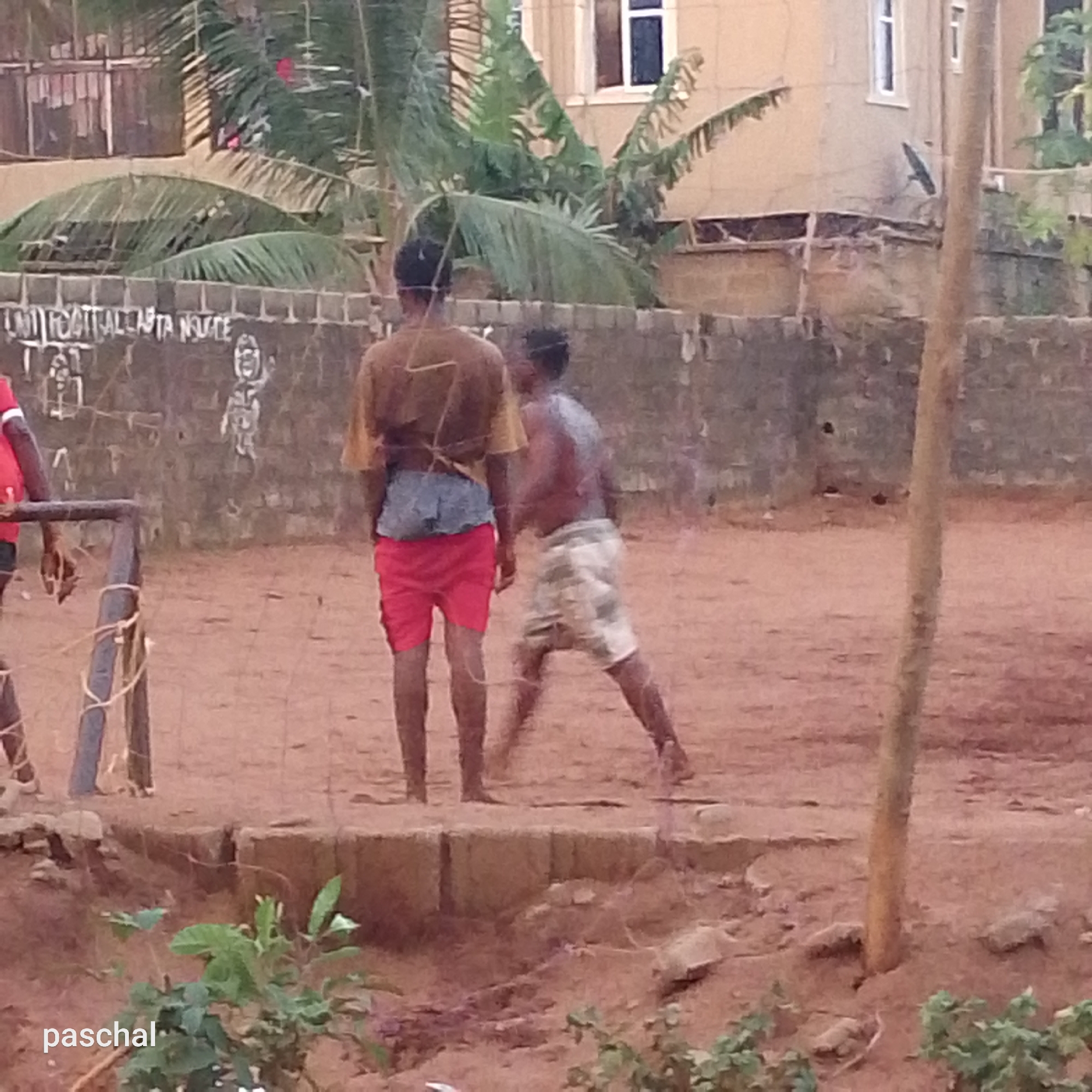 The image shows a sandy area in what appears to be a residential neighborhood, possibly in Ukwu-Abwa, Nigeria. Two males are the main subjects. One is wearing a tan shirt, grey shorts, and red shorts; he is facing away from the camera. The other male is shirtless and wearing patterned shorts. They are positioned near a low concrete structure. A section of a crude football goal is visible on the left. A low concrete wall with graffiti is in the background, along with houses and trees. The ground is bare earth. The word "paschal" is at the bottom left of the image.