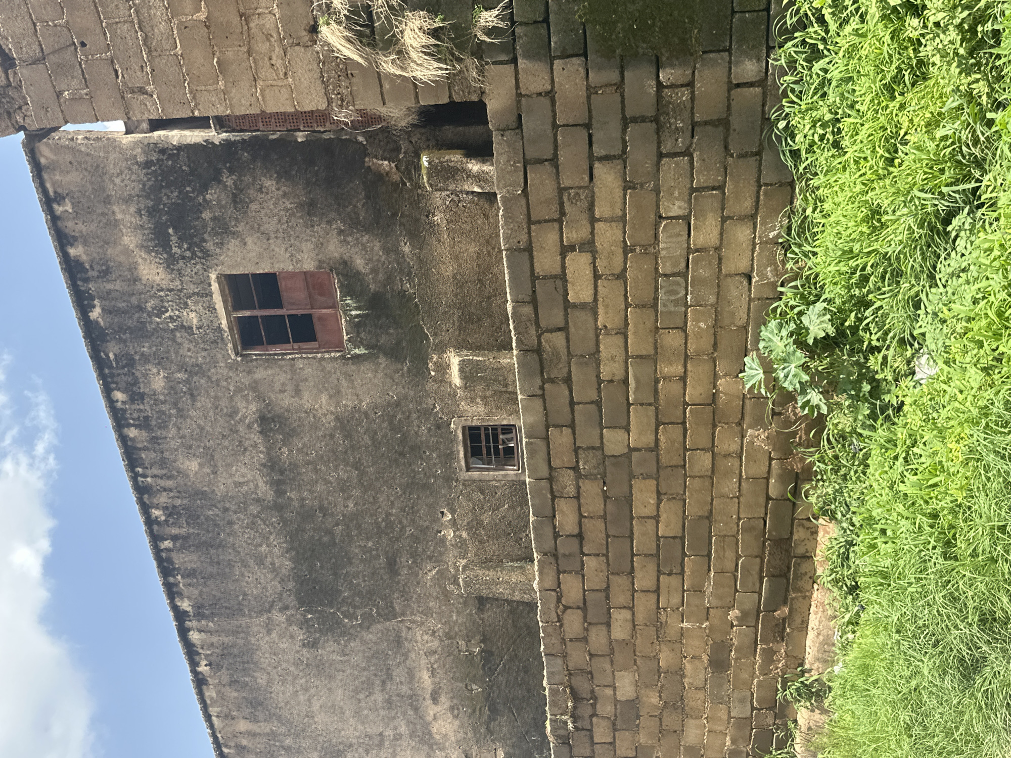 This media file shows an exterior view of an old, weathered building made of roughly textured grey concrete and brick. The building has at least two small windows with what appears to be dark, possibly metal, frames. One window is open, revealing a dark interior. The building is adjacent to a wall constructed of light brown bricks. To the right of the brick wall, there is an abundance of vibrant green grass and vegetation, suggesting an outdoor setting. The sky, visible in the upper left corner, is a clear blue with some wispy white clouds, indicating it is daytime and likely fair weather. The building's aged appearance, with stains and uneven surfaces, hints at its age and exposure to the elements. The scene appears to be in a rural or semi-rural environment, given the natural vegetation bordering the man-made structure. There is no visible text or people in the image. The overall impression is one of a quiet, possibly abandoned or neglected, historical structure amidst nature.