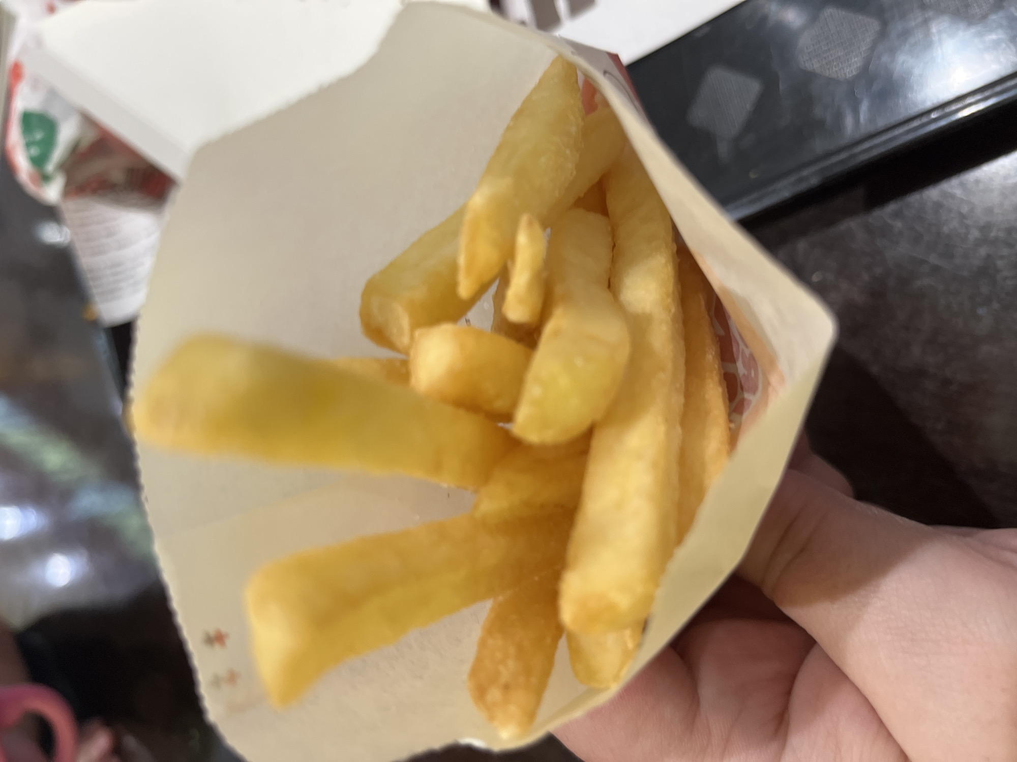 A close-up shot shows a paper bag filled with golden-brown french fries. A person's hand is holding the bag. The background is partially visible and shows a dark surface that might be a table. There is a hint of other objects or containers, but they are out of focus. The location of the image is not discernible from the visual content, thus not possible to confirm if it is in Sochi, Russia.