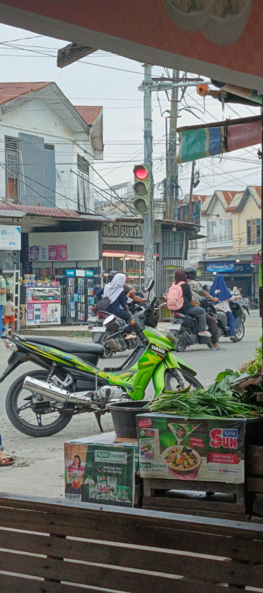 The image depicts a street scene in Garut, Indonesia, focusing on a busy intersection with motorbikes, pedestrians, and commercial storefronts. A prominent traffic light is visible, showing a red signal. Several motorbikes are on the road, some with riders. One motorbike in the foreground is bright green with "YAMAHA" branding. In the background, buildings line the street, including what appears to be a "NAR SURTA" establishment. Signs for "BSI" (Bank Syariah Indonesia) and "Hannochs IPS" are also visible. Various goods, including what look like packaged food products and produce, are displayed at a roadside stall in the lower right portion of the image. The overall atmosphere suggests an active urban environment during daylight hours, with a mix of traffic and commerce.