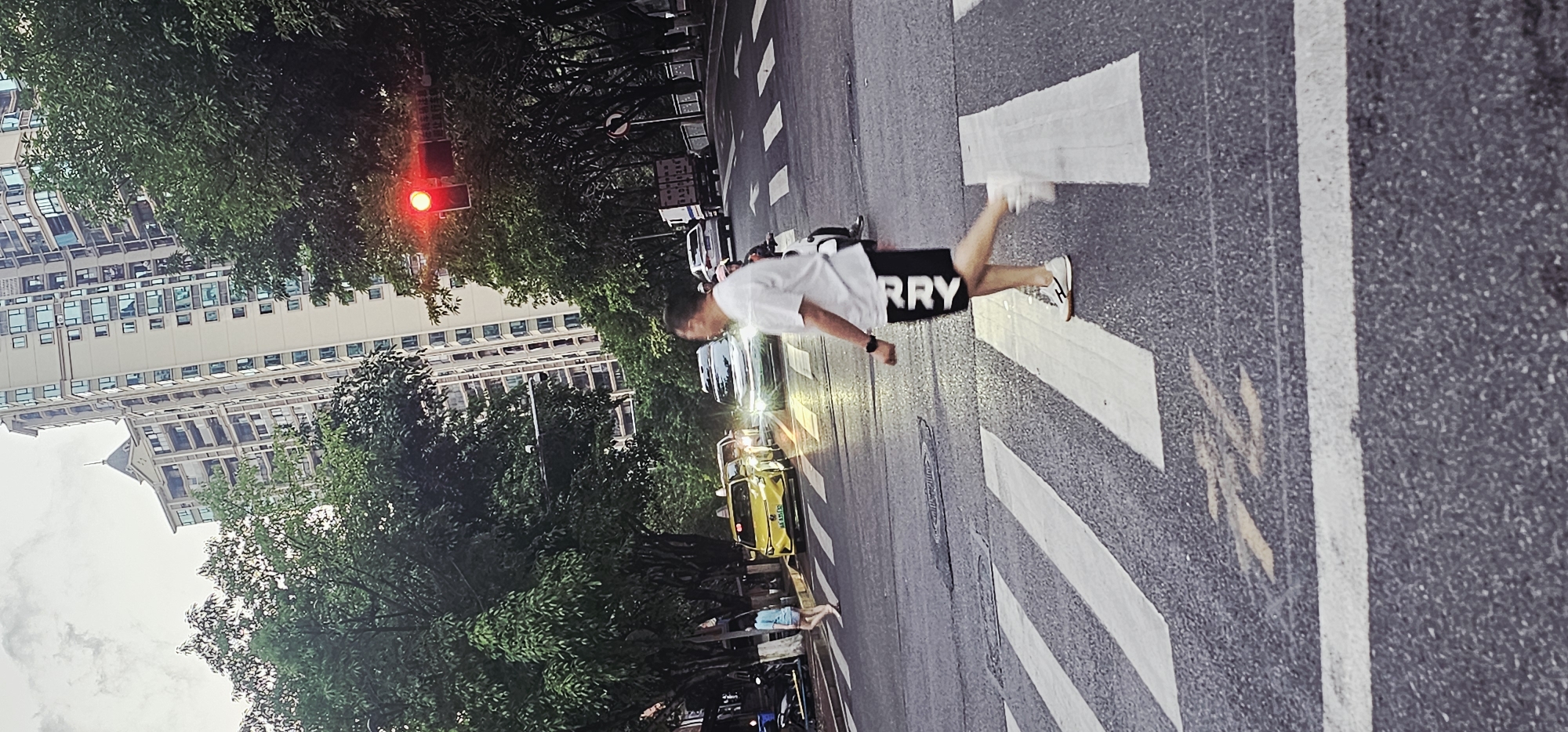 A pedestrian is crossing a crosswalk in Yuzhong District, Chongqing, China. The individual, wearing a white shirt and black shorts with white lettering, is mid-stride. Buildings and trees line the background and the traffic signal is showing red. Various vehicles, including a yellow taxi, are visible on the roadway.