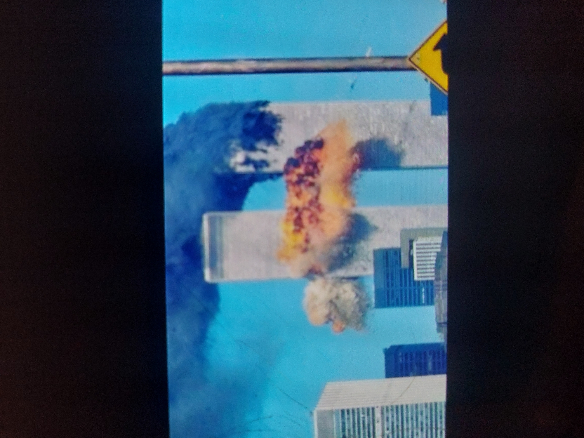 The image is a photograph of a screen displaying a catastrophic event involving the World Trade Center Twin Towers. The two prominent skyscrapers are depicted horizontally, suggesting the original image or screen display was rotated. A massive, fiery explosion, characterized by bright orange and red flames and thick dark smoke, erupts from the upper section of one of the towers. Below this primary blast, a smaller, lighter cloud of debris or smoke is also visible emanating from the same building. Dense plumes of smoke billow from the left side of the affected tower, obscuring parts of the structure and extending into the background.
The scene is set against a clear, bright blue sky, indicating daytime conditions. In the background and foreground, other city buildings can be seen, providing context to the urban environment. A horizontal metal bar, possibly part of a signpost or overhead structure, is visible in the upper part of the image. Adjacent to it, a portion of a yellow diamond-shaped sign with a black arrow pointing upward (or left, due to rotation) is present.
The image itself exhibits characteristics of a photo taken of a digital screen, including visible pixelation and slight glare. Black bars frame the left and right sides of the depicted scene, suggesting the original media's aspect ratio or display settings. No people are visible. The event depicted is clearly the attack on the World Trade Center on September 11, 2001.