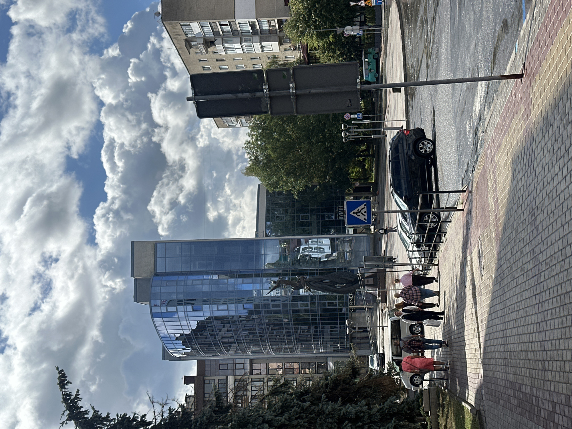 The image depicts a street scene in Ternopil, Ukraine. In the foreground, a paved sidewalk runs diagonally from the bottom left to the middle right of the frame, featuring a pattern of brickwork with alternating red and yellow sections. Several people are walking on the sidewalk. A group of five individuals, appearing to be adults, are walking away from the camera towards the right. One person is wearing a pink dress, another a plaid shirt, and another a dark vest.
Beyond the sidewalk is a street with several vehicles. A black SUV is parked or driving on the right side of the road. A white van is visible on the left side of the street, behind a group of people. In the background, there are modern glass-fronted buildings, one of which is a curved glass skyscraper reflecting the sky and surrounding structures. There are also older, multi-story apartment buildings with balconies. Trees line parts of the street and sidewalk.
Traffic signs are visible, including a pedestrian crossing sign with a blue background and a white silhouette of a person walking. The sky is partly cloudy with dramatic cumulus clouds and patches of blue sky, suggesting daytime. The overall impression is an urban environment with a mix of contemporary and older architecture.