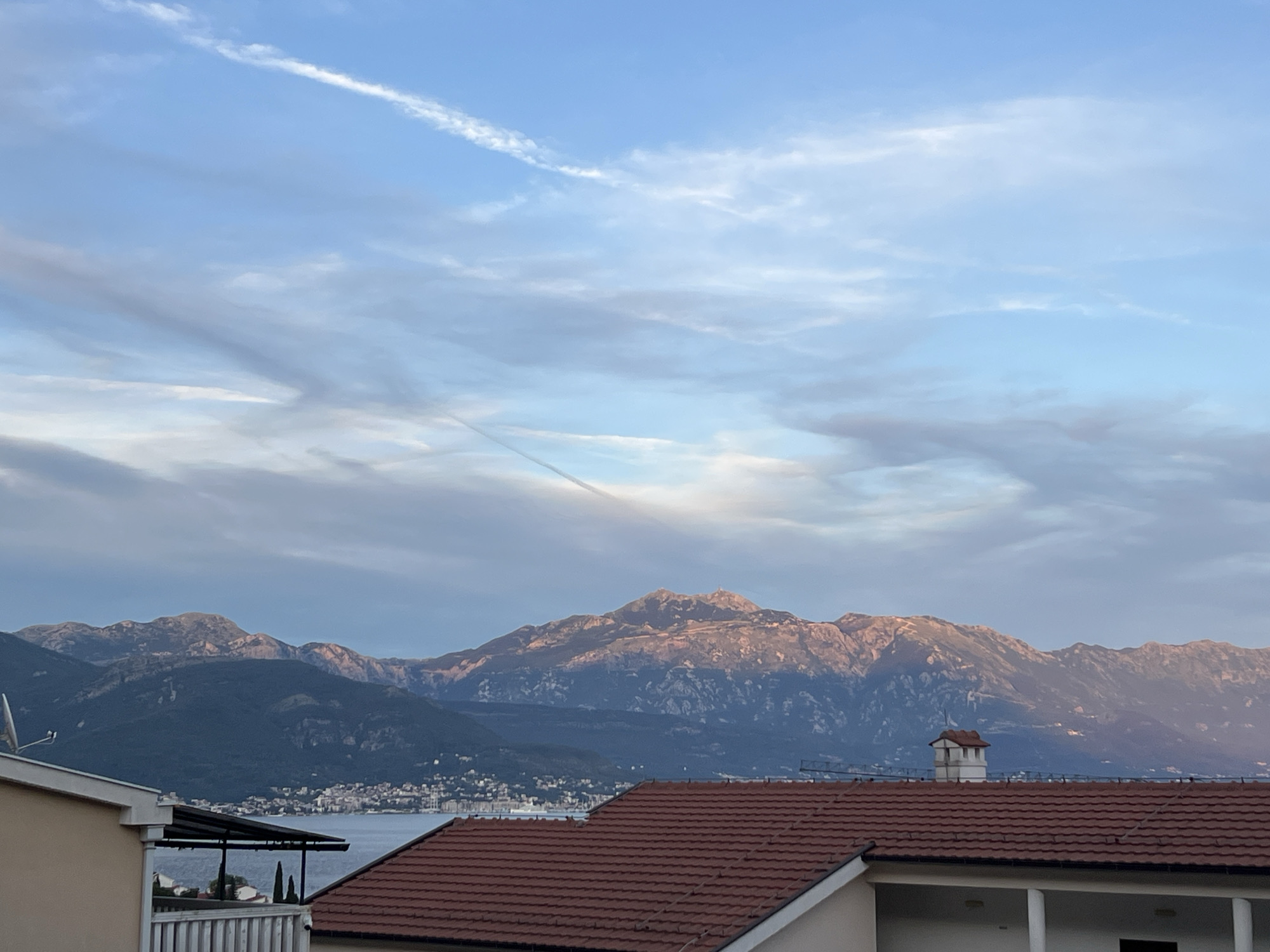 The image captures a panoramic view of the Bay of Kotor from Baošići, Montenegro. In the foreground, the terracotta tiled roof of a house is prominent, with the top of another building and a satellite dish visible to the left. A small, white building with a brown roof, resembling a chimney structure, stands on the rooftop in the middle ground. Beyond the buildings, the calm, blue waters of the bay stretch out, with a distant coastline dotted with a town and buildings. The backdrop is dominated by a majestic mountain range, its peaks illuminated by the warm, golden light of what appears to be either sunrise or sunset. The sky above is a gradient of soft blues and grays, with wispy clouds and a distinct contrail streaking across the upper portion. The scene is serene and picturesque, showcasing the natural beauty of the Montenegrin coastline.