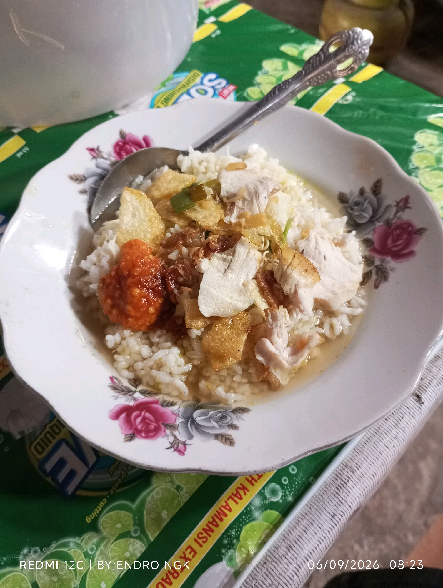 A close-up, overhead shot shows a white bowl of rice with shredded chicken, crispy fried onions, potato crisps, and a dollop of chili paste, served with a spoon. The bowl is placed on a green tablecloth adorned with lime slices and text, with a white pitcher and part of a yellow container visible in the background. The bottom of the image displays a timestamp and "REDMI 12C | BY: ENDRO NGK," suggesting the photo was taken in Ringinanom, Indonesia.