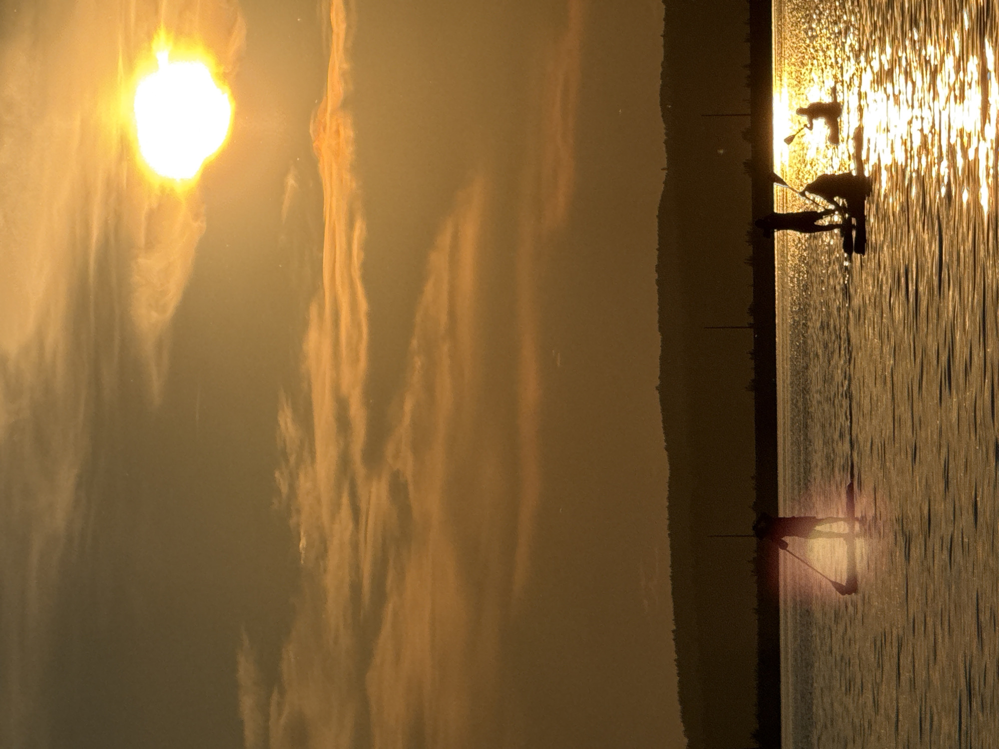 The image depicts a serene sunset over a body of water, likely a reservoir or river, in Chaikovsky, Russia. Silhouetted figures on paddleboards are visible on the water, enjoying the evening light. The sun, a bright orb, casts a warm golden glow, reflecting off the water's surface and illuminating the sky with hues of orange and yellow. Wispy clouds are scattered across the sky, adding texture to the scene. In the background, a dark, silhouetted shoreline with rolling hills suggests the natural landscape surrounding the city. The overall atmosphere is one of tranquility and peaceful recreation.
