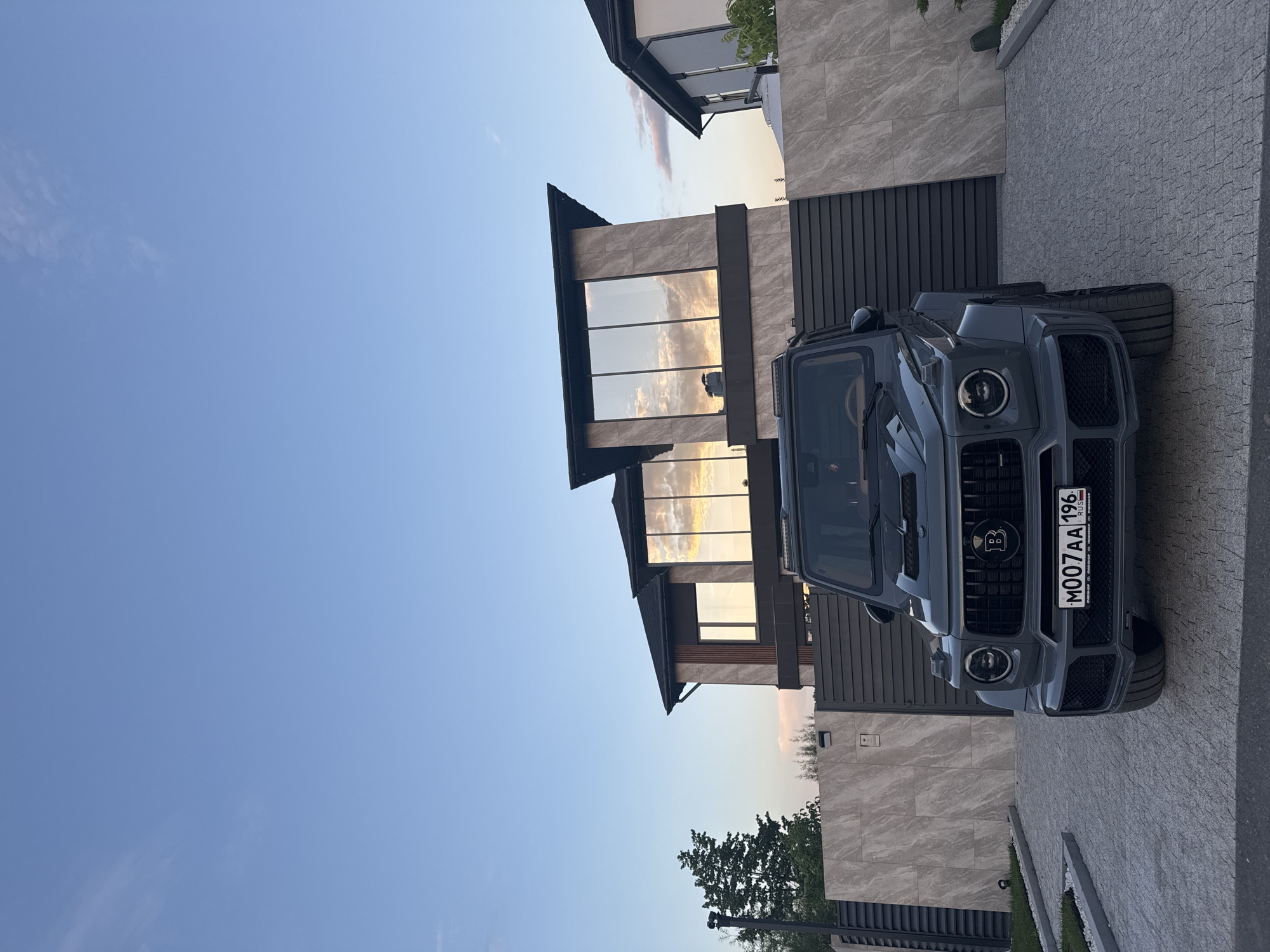 The media file captures a gray Mercedes-Benz G-Class SUV parked in a driveway next to a modern house. The house features large windows reflecting the sky, and is clad in light-colored stone and dark paneling. The sky is clear and blue, with a hint of sunset colors visible in the reflections on the windows. The time of day appears to be late afternoon or early evening. The setting is residential, likely a suburban or upscale neighborhood. There are no people visible in the image.
Notable details include the distinct shape of the G-Class, a luxury SUV. The car's front grille has a prominent "B" logo, indicating it is a Brabus model, a tuning company for Mercedes-Benz vehicles. The license plate is visible and reads "M007AA 196 RUS". The overall mood is one of luxury and tranquility. The location is identified as Yekaterinburg, Russia.