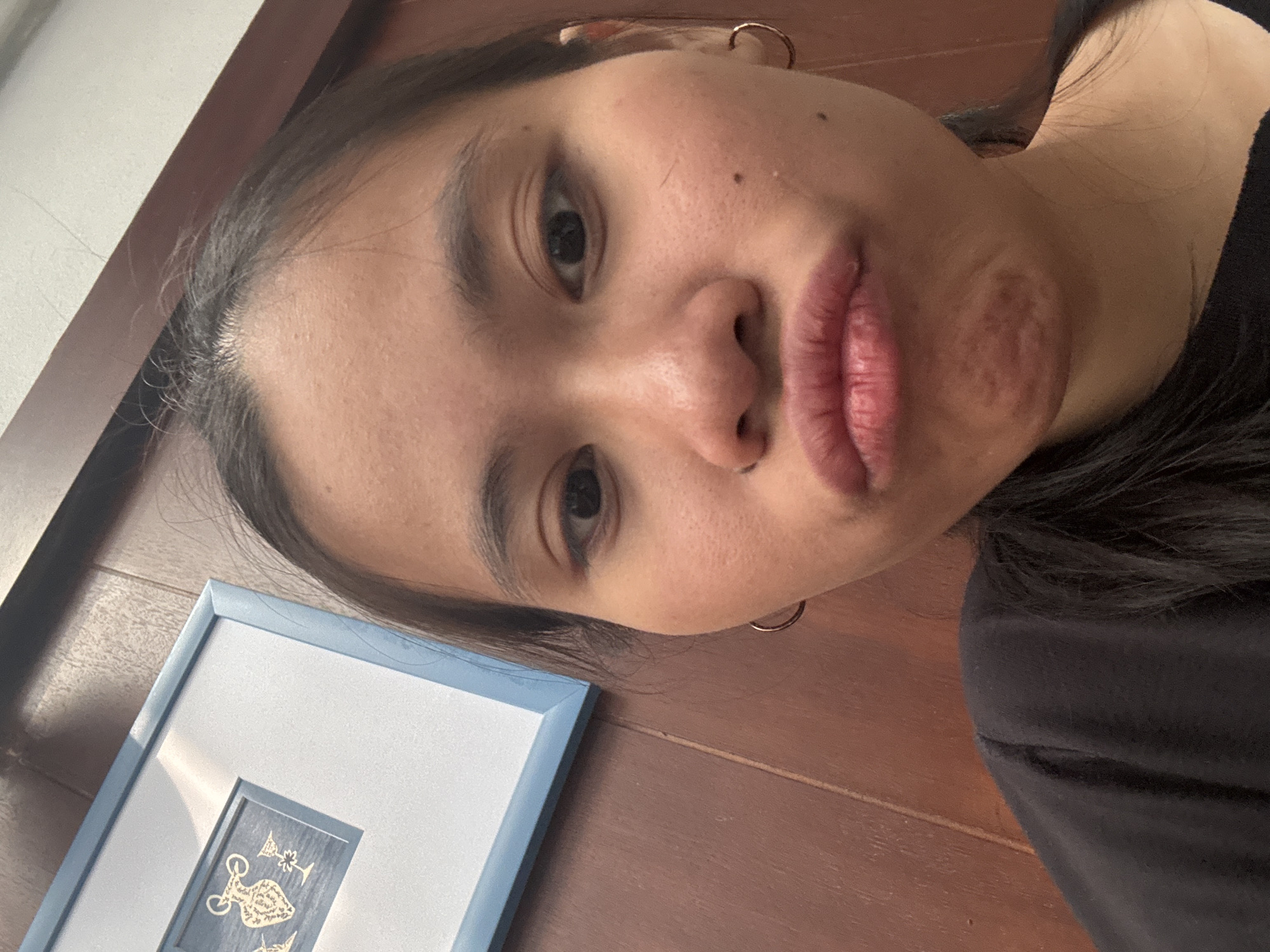 An upper-body, eye-level, medium close-up shot shows a young person of Asian descent looking directly at the camera in a selfie. The person has dark hair pulled back, dark eyes, and is wearing a black t-shirt and small hoop earrings. Their mouth is pursed into a "fish face" pose. The background consists of a wooden paneled wall, and to the left, a framed picture with a blue border and a white matte. The picture contains a dark blue inset with a gold decorative design of what appears to be amphorae and goblets. The lighting is natural and appears to be coming from above and to the left, casting slight shadows. The scene appears to be indoors, possibly in a residential setting in South Jakarta, Indonesia.