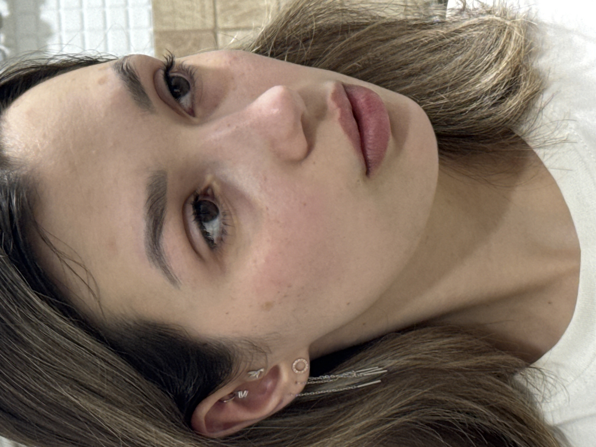 This is a close-up, rotated portrait of a person, likely a young woman, lying on their left side. Their face is angled towards the viewer, with their gaze directed slightly upwards and to the right. The person has fair skin with a few scattered freckles or beauty marks, particularly visible on the cheek and forehead. Their lips are full and appear to have a soft pink tint, possibly from lip balm or a natural shade. Well-defined, dark eyebrows frame their eyes, which are open and dark in color with long, dark eyelashes.
The person's medium brown hair, with lighter highlights, frames their face and extends onto a white surface, possibly a pillow or bed sheet. To the right of the image, the person wears a white garment, likely a t-shirt, visible at the neckline. Their visible right ear is adorned with multiple silver-toned piercings: a hoop-like piercing on the tragus, another small piercing on the helix, and two earrings on the lobe – one a circular stud and the other a dangling chain earring featuring multiple small bar-like elements. The background directly behind the person's head appears to be a light-colored tiled wall. The lighting is soft and even, highlighting the contours of the face without harsh shadows. No text is visible in the image. The expression is neutral and calm.