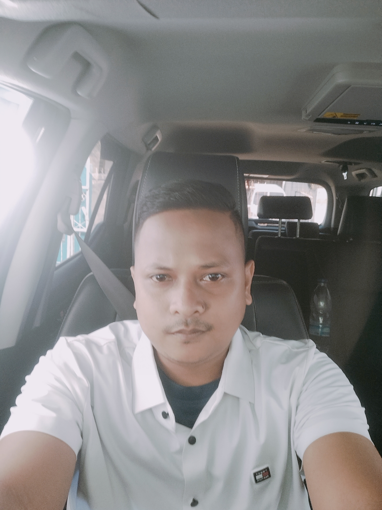 A man with short, dark hair and a mustache is sitting in the driver's seat of a car. He is wearing a white collared shirt over a dark t-shirt. The car's interior is visible, including the steering wheel, dashboard, and headrests of the seats. The man is looking directly at the camera. Outside the car, a street scene is visible, with buildings and other vehicles. The image appears to have been taken during the day, with sunlight streaming into the car. The location is identified as Lhokseumawe, Indonesia.