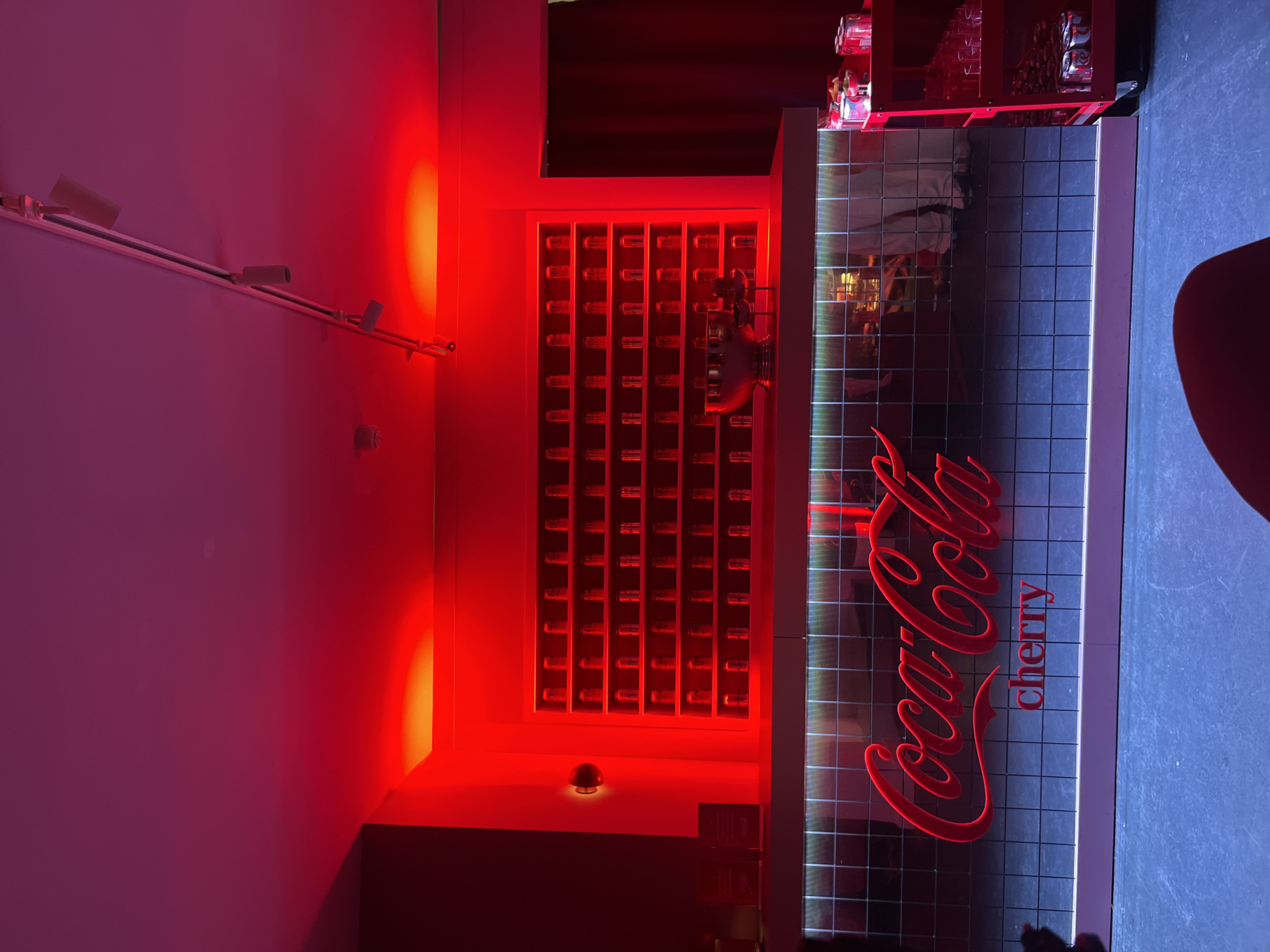 This media file captures an indoor setting, likely a themed bar or lounge, characterized by striking neon and ambient lighting. No people are visible, suggesting the space is unoccupied or awaiting guests.
The central focal point is a prominent red neon sign on the front of a bar or counter, clearly displaying the text "Coca-Cola" in its iconic script, with "cherry" written below it. The surface of this bar/counter features a modern, illuminated grid pattern.
To the left of the bar, a wall is dramatically illuminated by a bright red spotlight, casting a strong red glow. Below this, there is a display unit with multiple compartments, each holding small, cylindrical, translucent objects, possibly bottles or vials, which are also glowing with a reddish hue. A track lighting fixture with several white light heads runs horizontally along the upper part of this wall, which transitions from red to a magenta or purplish tone further left.
The floor or a lower section on the right side of the image appears dark blue, contrasting with the red lighting. The overall aesthetic is modern and vibrant, creating an energetic atmosphere. The strong branding suggests a commercial or promotional space, possibly for a special event or a dedicated Coca-Cola themed venue in London, United Kingdom. The lighting conditions imply a night-time setting or an interior with controlled artificial light.
