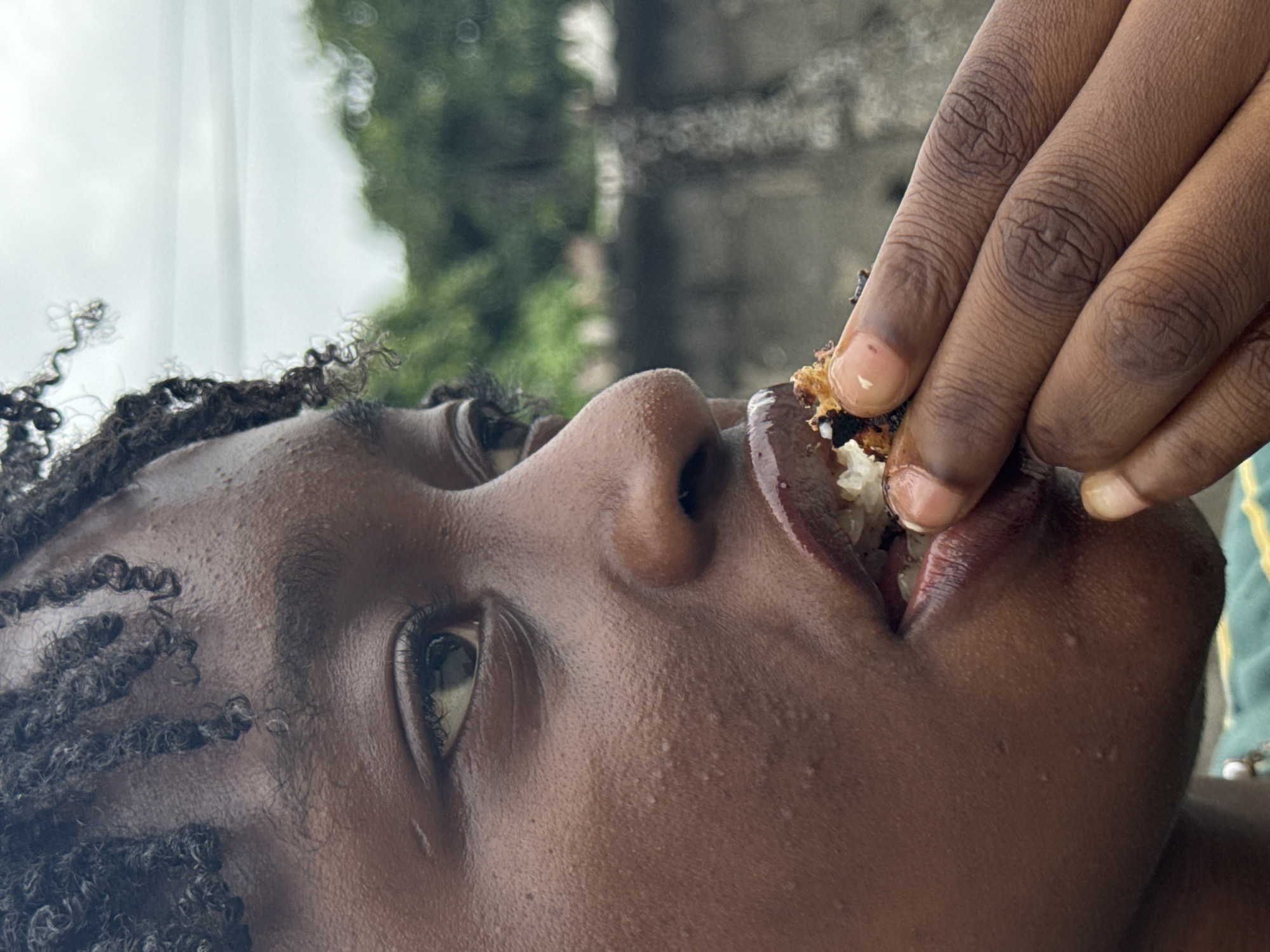 The image depicts a close-up, tilted view of a person with dark skin and curly hair being fed. A hand with dark skin is holding a piece of food, composed of small, white rice-like grains and a darker, perhaps charred, component, and is positioned near the person's mouth. The person is looking upwards. The background is blurry, showing green foliage and a grey structure possibly indicative of a building or wall.
Specific location details within Ado, Nigeria cannot be determined from the image alone.
