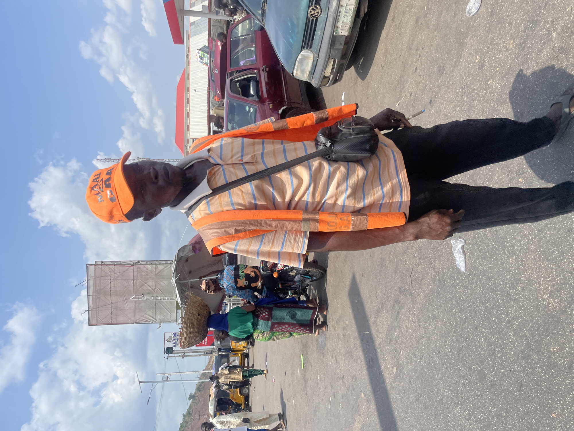 The image depicts a street scene in Lokoja, Nigeria. An older man, wearing an orange cap and vest over a striped shirt, stands near a road, holding what appears to be a pen. He has a small black bag across his body. Behind him, traffic is visible, including a dark red car and a green Volkswagen. Further down the road, there's a woman carrying a basket on her head, surrounded by other people and vehicles, including yellow tricycles (keke napep). A large advertising structure is in the background. The road surface is paved. The sky is blue with scattered clouds.