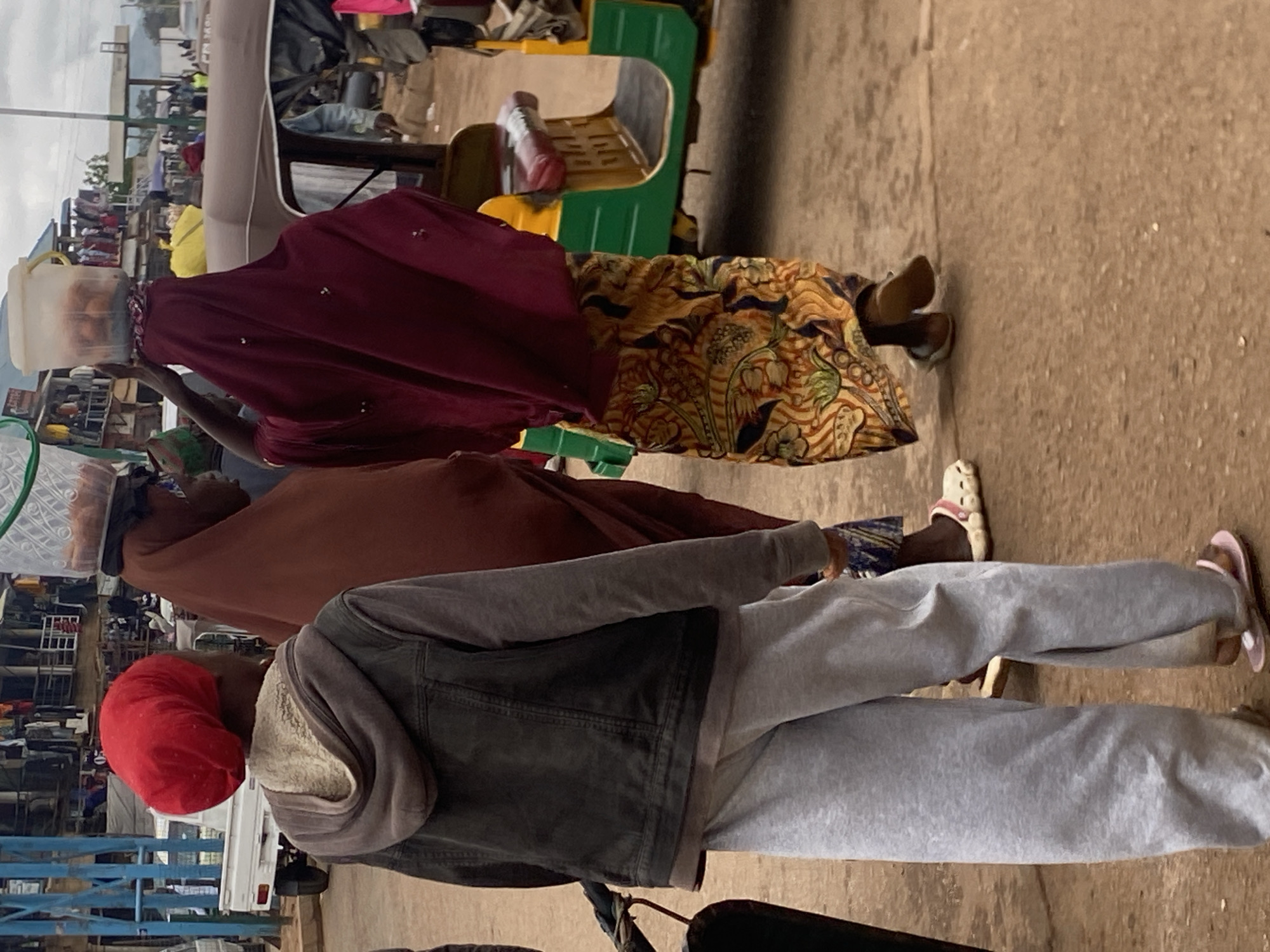 In Bukuru, Nigeria, a street scene captures individuals navigating a marketplace. In the foreground, a person wearing a red headscarf, a grey hoodie layered under a dark denim jacket, and light grey sweatpants walks away from the camera. To their right, another individual, wearing a deep red head covering and a flowing maroon robe, balances a clear plastic container filled with brown items on their head. Their patterned skirt is a vibrant blend of gold, green, and brown. Further back and to the left, a third person, partially obscured, wears a brown head covering. They are also carrying a similar clear plastic container on their head. A brightly colored auto-rickshaw, predominantly green and yellow with a brown canopy, is parked to the right of the frame, suggesting transportation is readily available. The ground is a dirt or gravel surface, common in many urban areas. In the background, stalls and structures indicative of a market are visible, with goods displayed and people moving about. The overall impression is one of a bustling commercial hub where everyday commerce takes place.