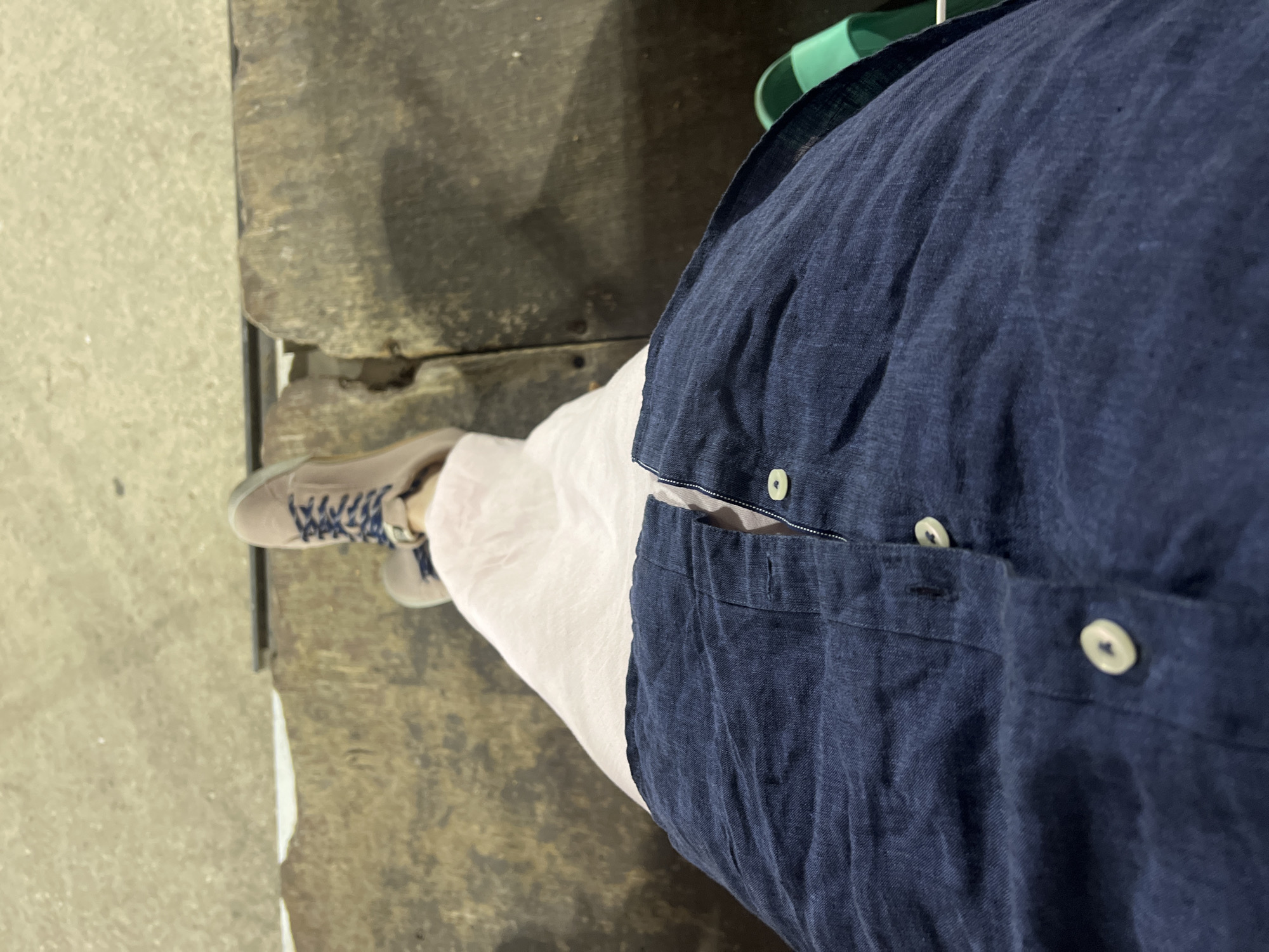 This is a first-person perspective, low-angle shot of a person sitting on a rough, weathered wooden bench. The person is wearing a navy blue button-down shirt that is open at the top, revealing a glimpse of a pink garment underneath. The shirt has two visible white buttons and a buttonhole. On the person's feet are light brown sneakers with dark blue laces. To the right, a bright green sandal is visible. The background appears to be an outdoor urban setting, possibly a boardwalk or promenade. The bench itself is dark and textured, with visible wear and tear. The ground in front of the bench is a light grey, uneven surface, possibly concrete or paving stones. The scene is dimly lit, suggesting it might be evening or an overcast day. The composition focuses on the lower half of the body and the immediate surroundings.