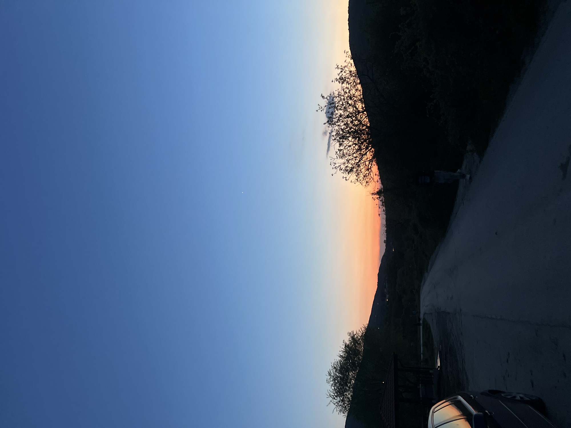 Captured in Galabovo, Bulgaria, the image presents a landscape at twilight. The sky transitions from a deep blue on the left to hues of orange and pink near the horizon. A dark, mountainous silhouette forms the backdrop along the right side, interspersed with sparse vegetation and a bare tree. A winding road occupies the right foreground, its surface dimly lit. A parked car is partially visible at the bottom edge of the frame.