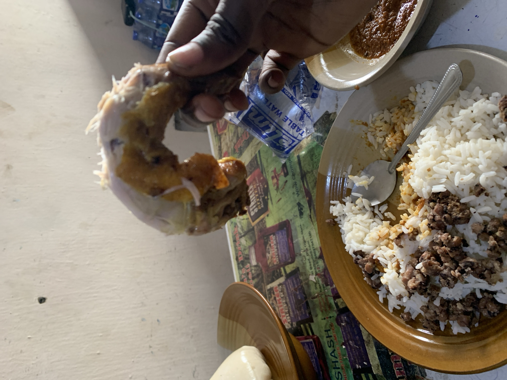 The image shows a hand holding a piece of cooked chicken with a bite taken out of it, next to a plate of rice with some meat on top and a bowl of sauce. The scene is set on a table with a printed tablecloth. A package of "Elum TABLE WATER" can be seen on the table. The tablecloth contains some text that states, "SHASH INDUSTRIES LIMITED," and "ai Road, Da Ogtin State." It also shows a contact phone number and email.