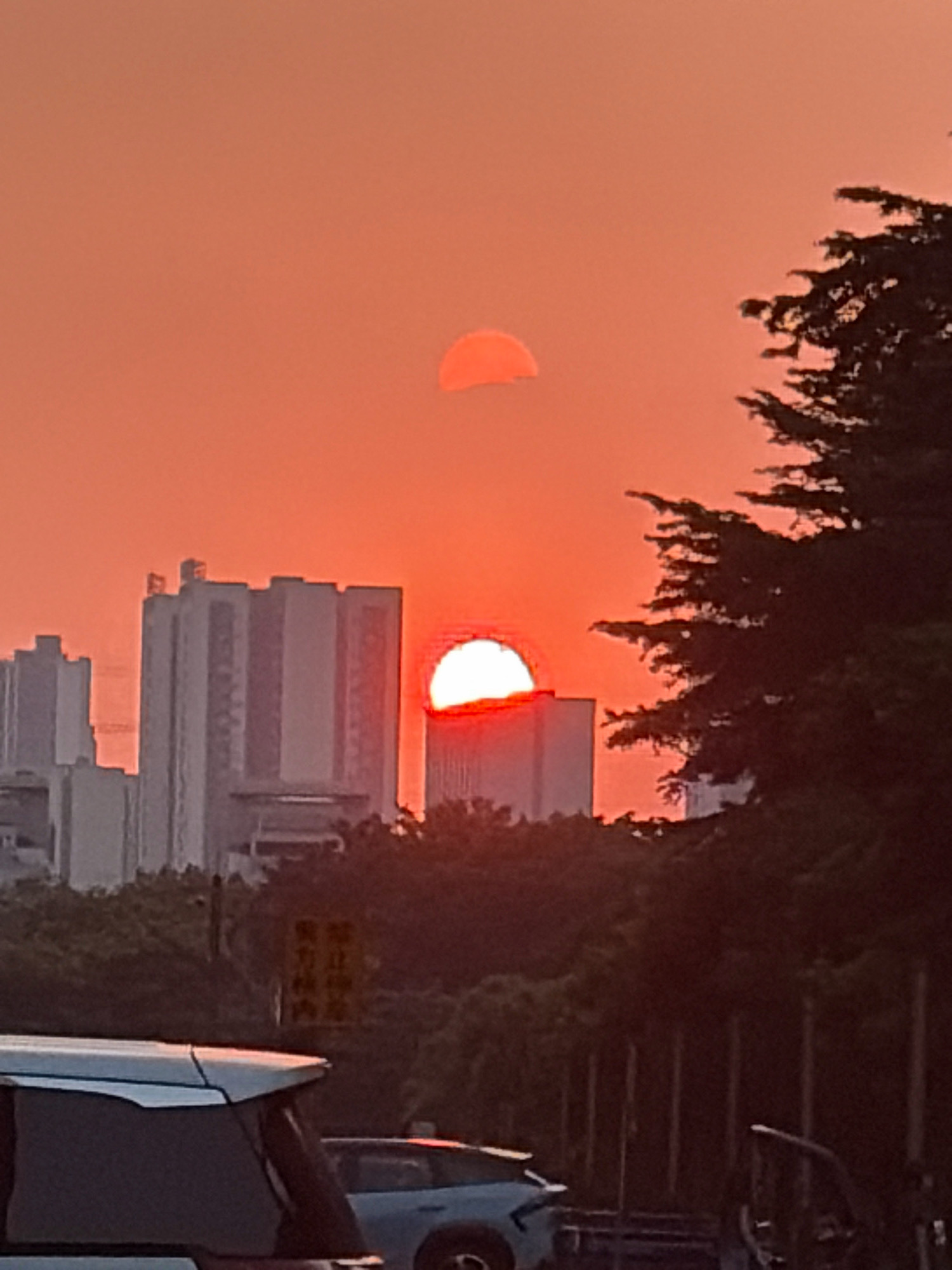 The image depicts a sunset scene in Chancheng District, China. Two suns are visible in the sky, one higher and smaller, and the other larger and partially obscured by buildings. Tall, rectangular buildings dominate the background, with dense foliage in the middle ground. In the foreground, the roof and rear windshield of a white vehicle are visible, along with a darker vehicle to its right. The sky is a vibrant orange, characteristic of a sunset.
