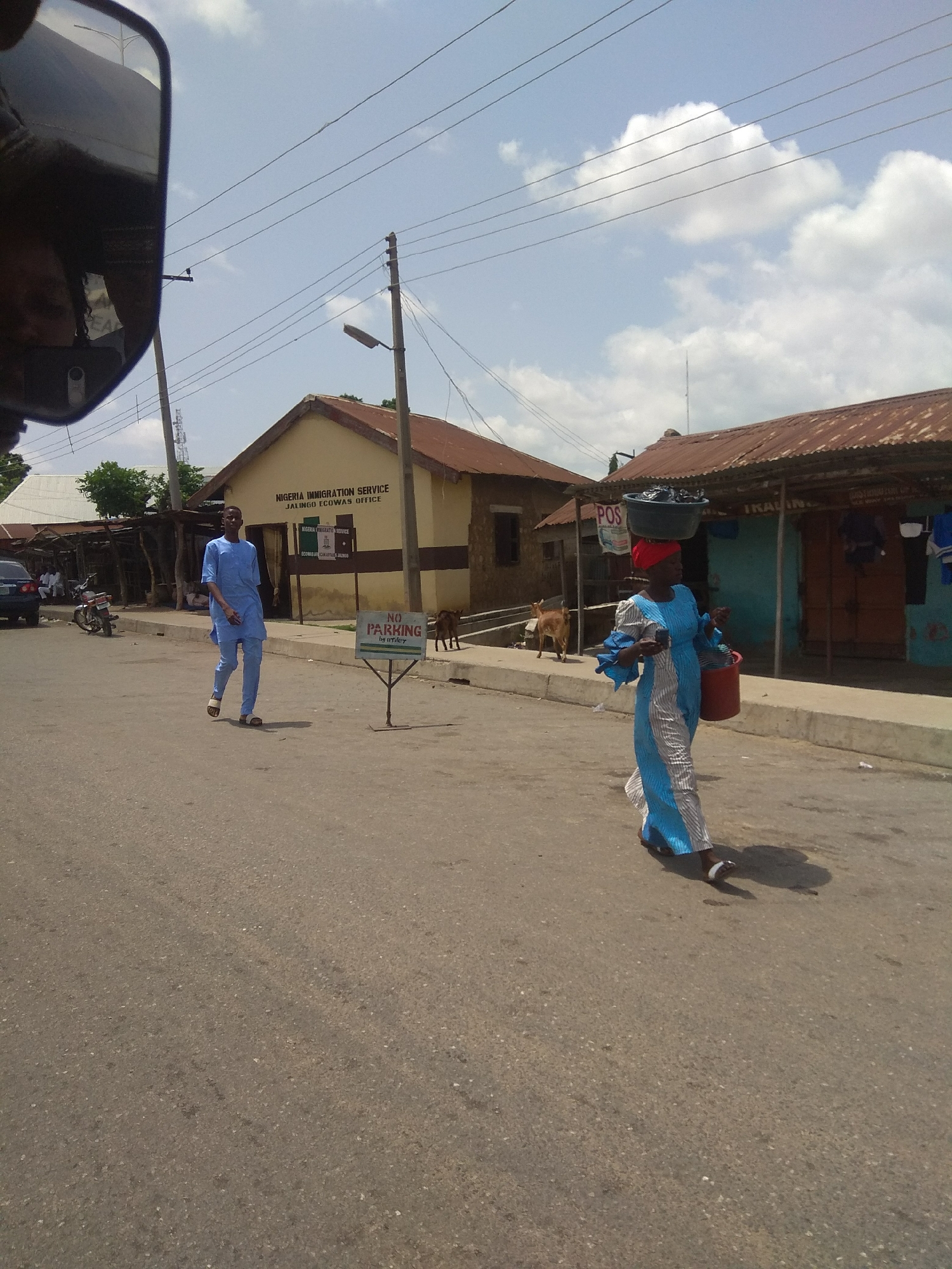 A medium shot captures a street scene in Jalingo, Nigeria, under a bright, partly cloudy sky. The foreground is dominated by an asphalt road. On the right side of the road, a woman walks towards the viewer, carrying a large basin filled with items on her head and a red bucket in her right hand. She wears a long, flowing blue and white striped dress and a red headscarf.
To the left of the woman, a man in a light blue traditional outfit walks away from the viewer, towards a building with "NIGERIA IMMIGRATION SERVICE JALINGO ECOWAS OFFICE" written on its facade. A "NO PARKING" sign stands in front of the immigration office. Further down the street, a goat is visible near the immigration building.
Behind the immigration office, another building with a corrugated metal roof suggests a commercial area, with "POS" and "IKALING" visible on signage. Various objects, including clothing, are displayed outside this establishment. In the background, more structures and trees are visible, along with power lines crisscrossing the sky. A motorcycle is parked to the left of the immigration building. The scene appears to be set during daylight hours, with a clear, sunny atmosphere.