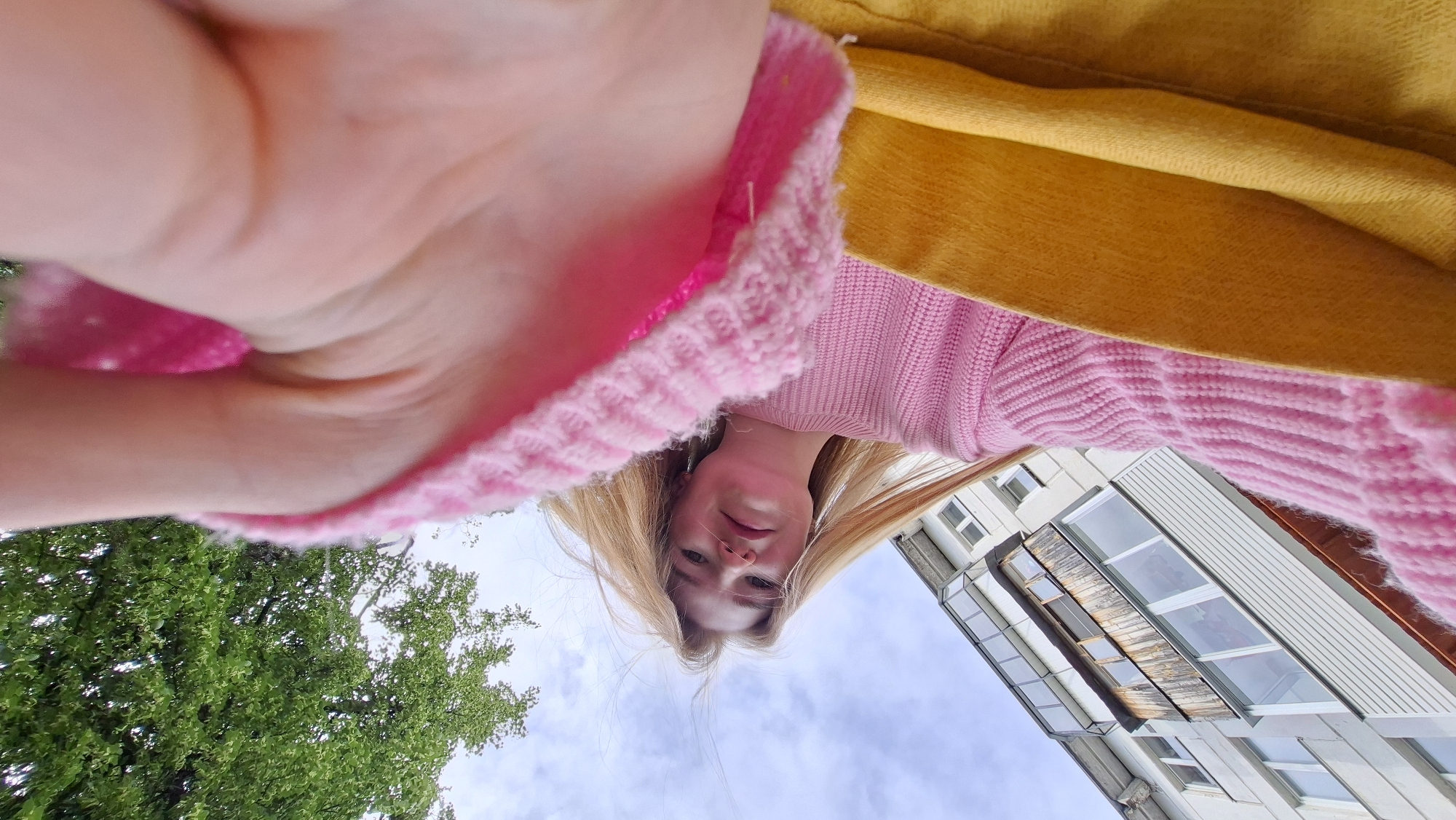 This is an unusual low-angle selfie featuring a young woman with long, straight blonde hair, looking directly upwards at the camera. She is wearing a pink knitted sweater, partly obscured by a bright yellow garment, possibly a coat or jacket. Her hand, holding the camera, is visible in the upper left corner.
The outdoor setting is captured from this unique perspective, revealing a partly cloudy daytime sky. To the left, lush green trees are visible, indicating a natural environment. To the right, a multi-story building with numerous windows and a distinctive bay window or balcony structure is prominent, suggesting an urban or suburban apartment complex.
The woman has a neutral to slightly content expression. There is no visible text in the image. The scene is set in Chaikovsky, Russia.