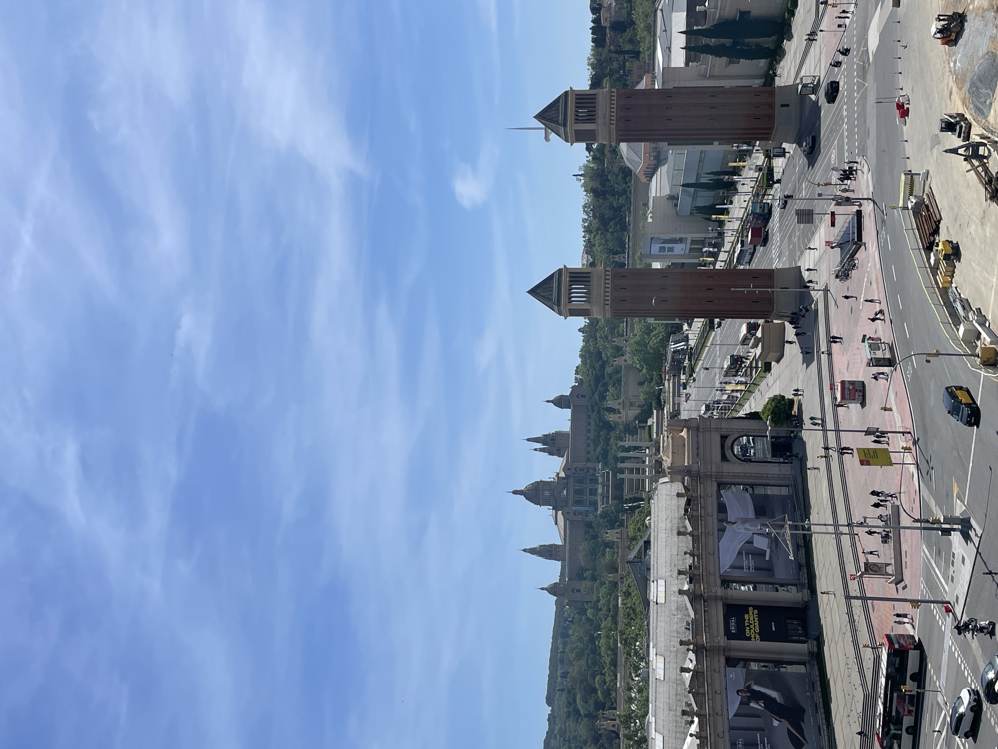 This high-angle, vertically oriented photograph captures a bustling scene from Plaça d'Espanya in Barcelona, Spain, under a clear, bright blue sky with light, wispy clouds. The sun illuminates the square, indicating midday or early afternoon.
In the midground, the two distinctive red-brick Venetian Towers (Torres Venecianes), with their green, conical roofs, stand as prominent architectural landmarks. Beyond them, positioned majestically atop a tree-covered hill, is the grand Palau Nacional (National Palace) of Montjuïc, featuring multiple domes and an impressive facade. The surrounding areas of Montjuïc are lush with greenery.
Below the towers, a wide, multi-lane road system crisscrosses the square, with a visible red bus, cars, and motorcycles moving along. Numerous pedestrians are active on the sidewalks and at crosswalks, suggesting a vibrant urban environment. To the right, some construction equipment and materials are present next to the road.
On a large building in the lower left, likely part of an exhibition center such as Fira de Barcelona, prominent advertising banners are displayed. Visible text on these banners includes "BRIDAL" and "ON THE SHOULDERS OF GIANTS," suggesting an event or exhibition is taking place. Streetlights, lampposts, and parked bicycles are also scattered throughout the pedestrian areas. The scene conveys a sense of dynamic city life amidst historic architecture.