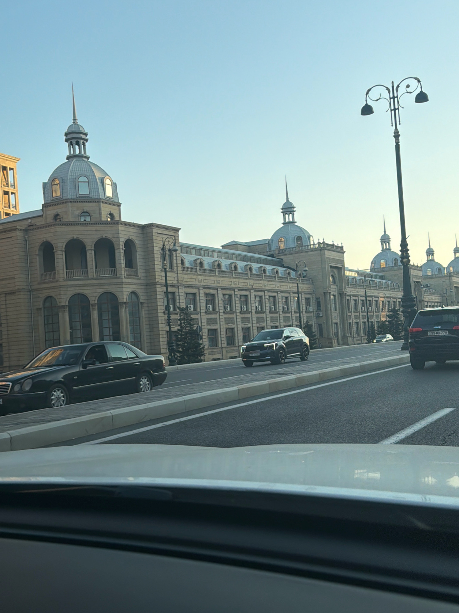 The image captures a street view in Baku, Azerbaijan, with a focus on a grand building and passing cars. The building, characterized by its classical architecture featuring domes, spires, and arched windows, likely houses a significant government or cultural institution. The sky is a clear, pale blue, suggesting it's either early morning or late afternoon. Several vehicles are visible on the road: a black Mercedes-Benz sedan in the foreground on the left, a modern black SUV in the middle of the road, and a black Jeep SUV on the right. A decorative street lamp with two lights stands prominently on the right side of the road, adding to the urban aesthetic. The road itself is paved and marked with white lines, indicating a well-maintained city thoroughfare. The perspective suggests the photo was taken from within a moving vehicle.