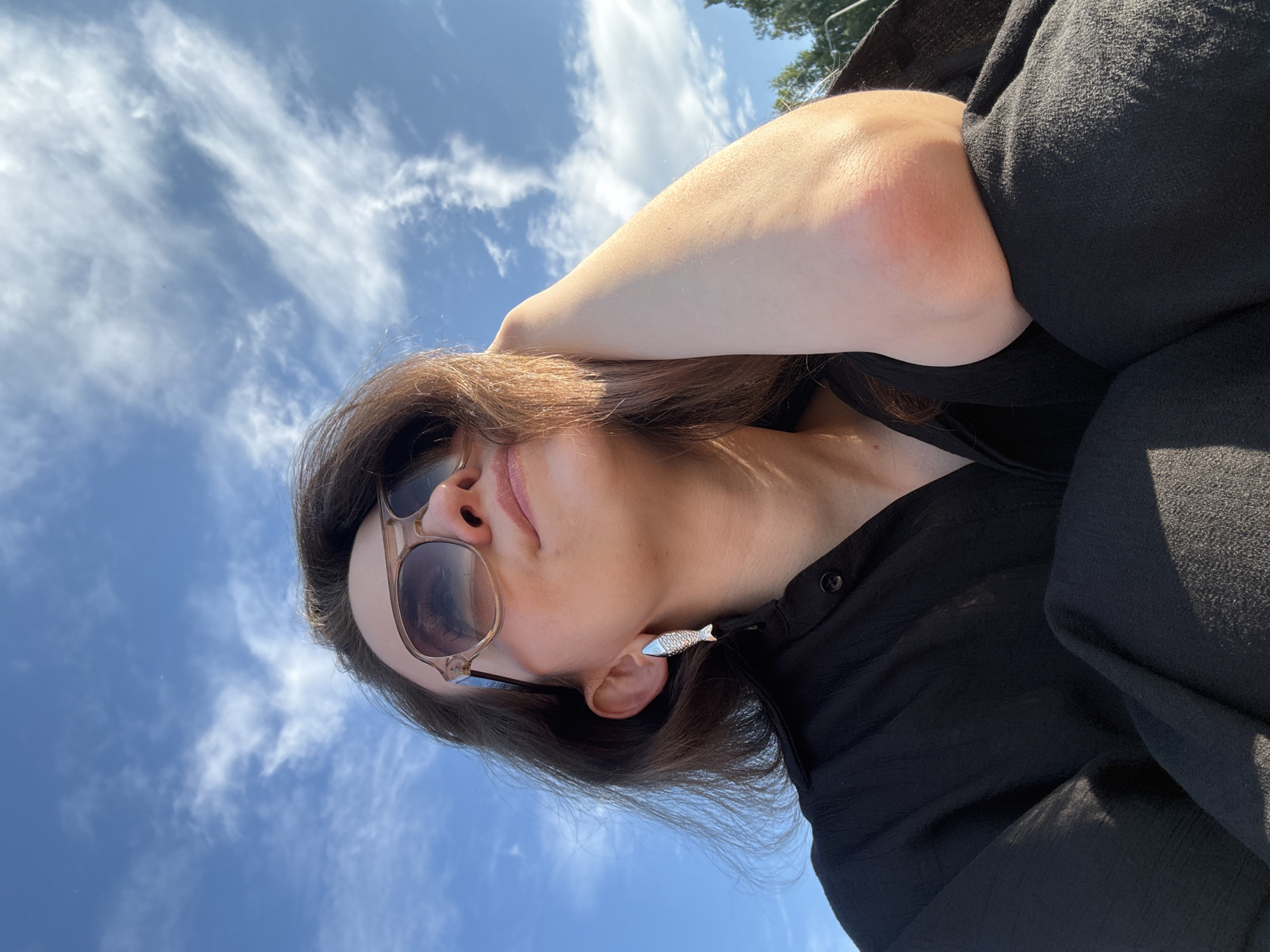 In an outdoor, low-angle shot against a bright blue sky with scattered white clouds, a woman is seen from the chest up, looking upwards and to the right. She has long, dark brown hair, wears large, light brown-framed sunglasses, and a black collared shirt. A fish-shaped silver earring dangles from her left ear. Her right arm is bent at the elbow, with her forearm resting against her forehead, and her elbow is visible. The background shows the sky, with a faint hint of green foliage on the right side. The image appears to be taken in Chaikovsky, Russia, on a sunny day.